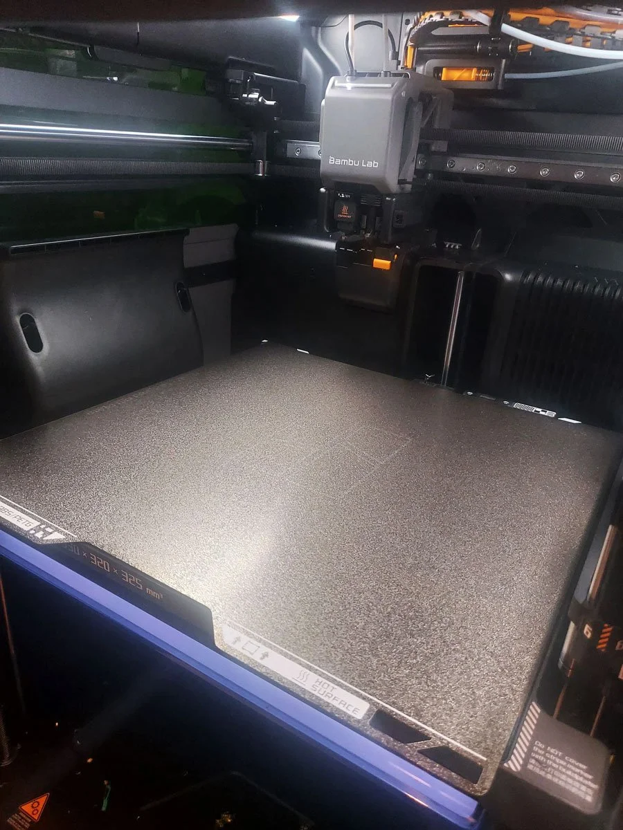 The interior of a 3D printer. The print bed is visible with a textured surface.