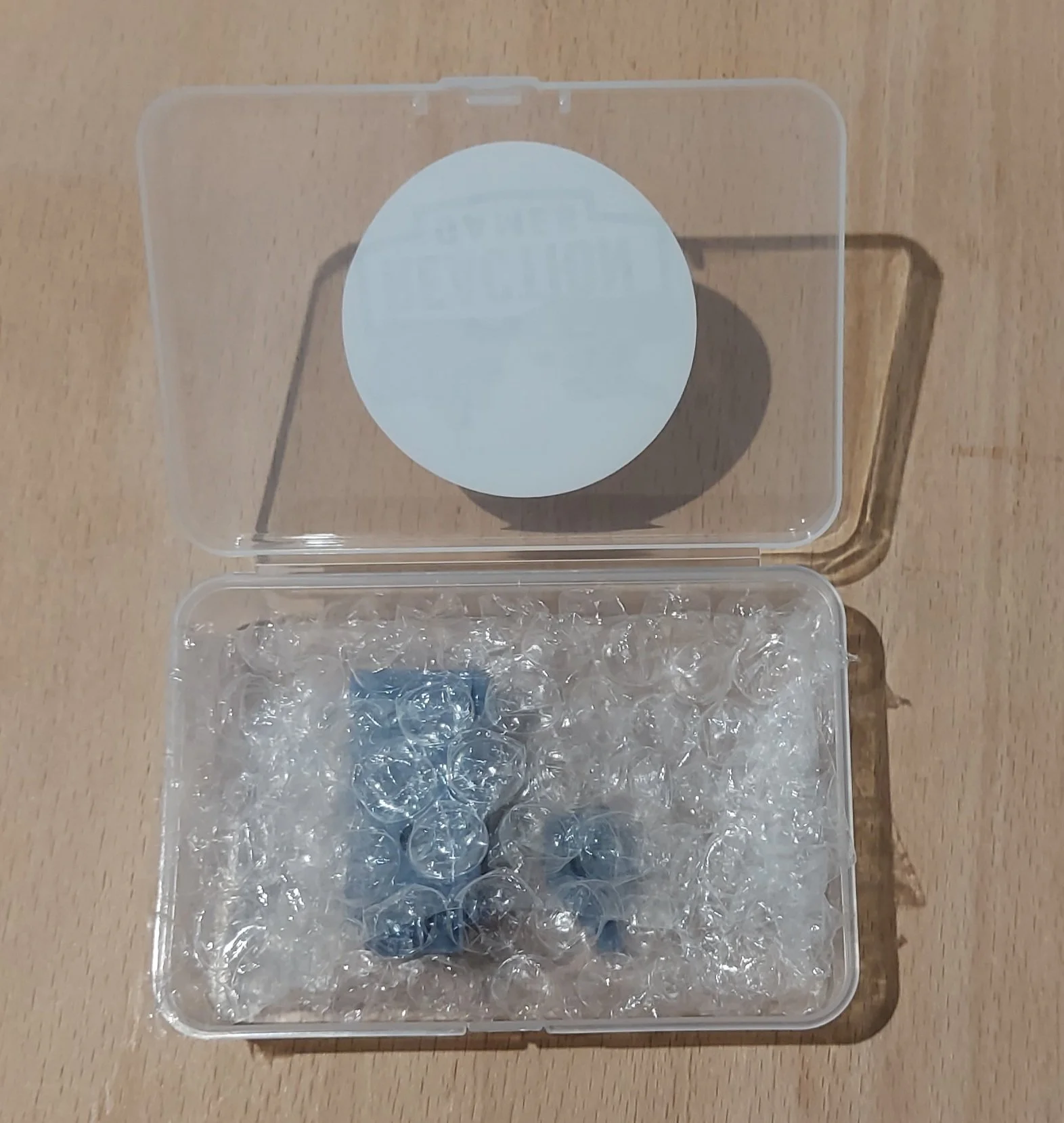 Open clear plastic box containing bubble wrap packaging and several small blue and silver items inside on a wooden surface.