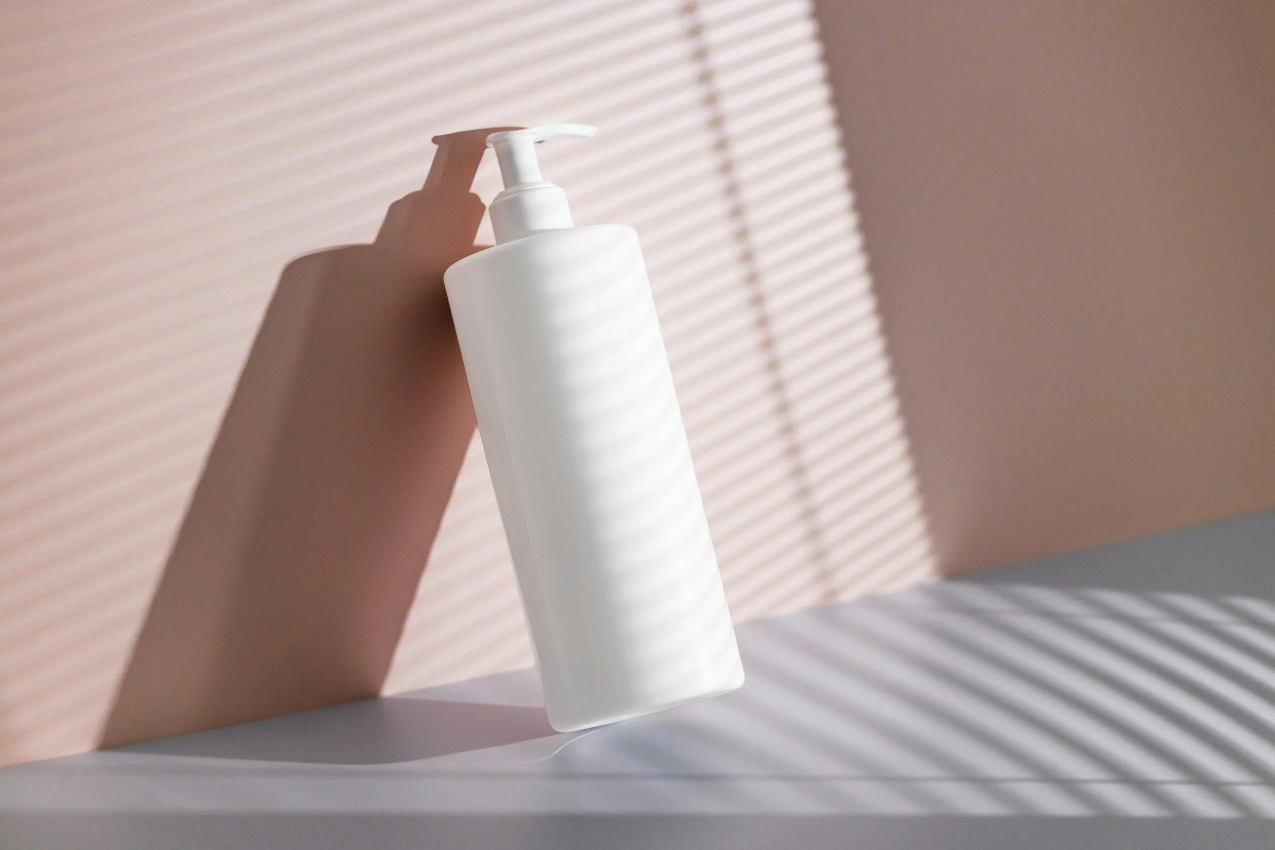 A white plastic pump bottle standing on a white surface with striped shadows and a blurred beige wall in the background.