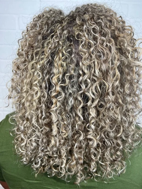 Back of a person with long, tight, blonde curly hair, wearing a green shirt.