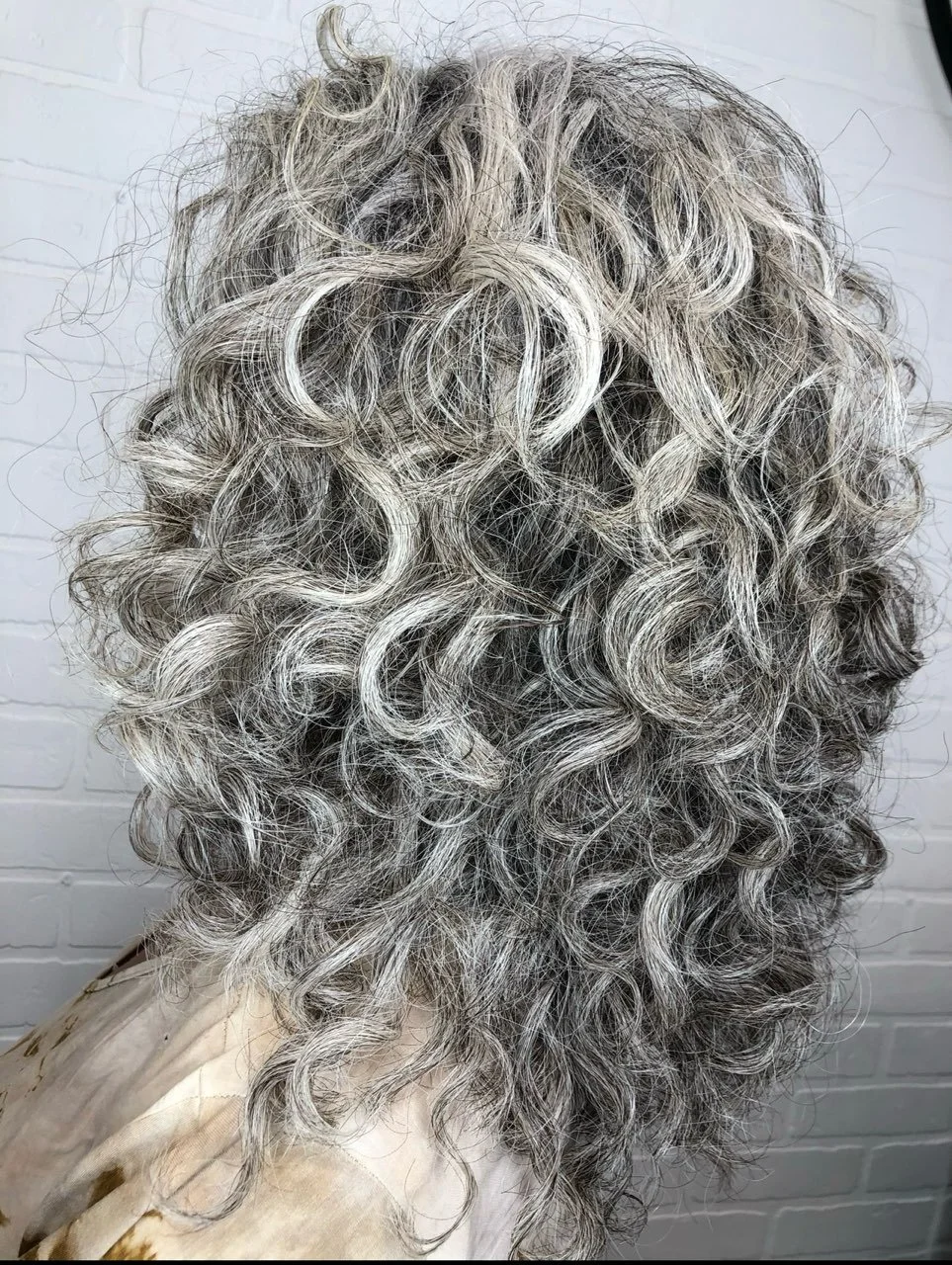 A close-up of curly gray hair on a person with a light-colored top, standing in front of a white brick wall.