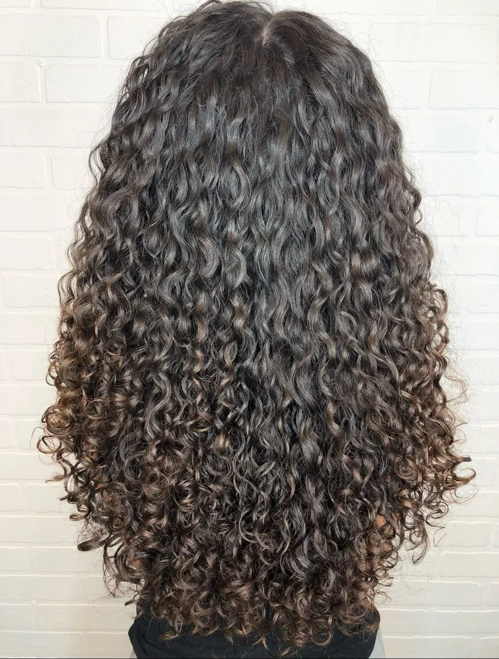The back view of a person with long, dark, curly hair against a white brick wall.