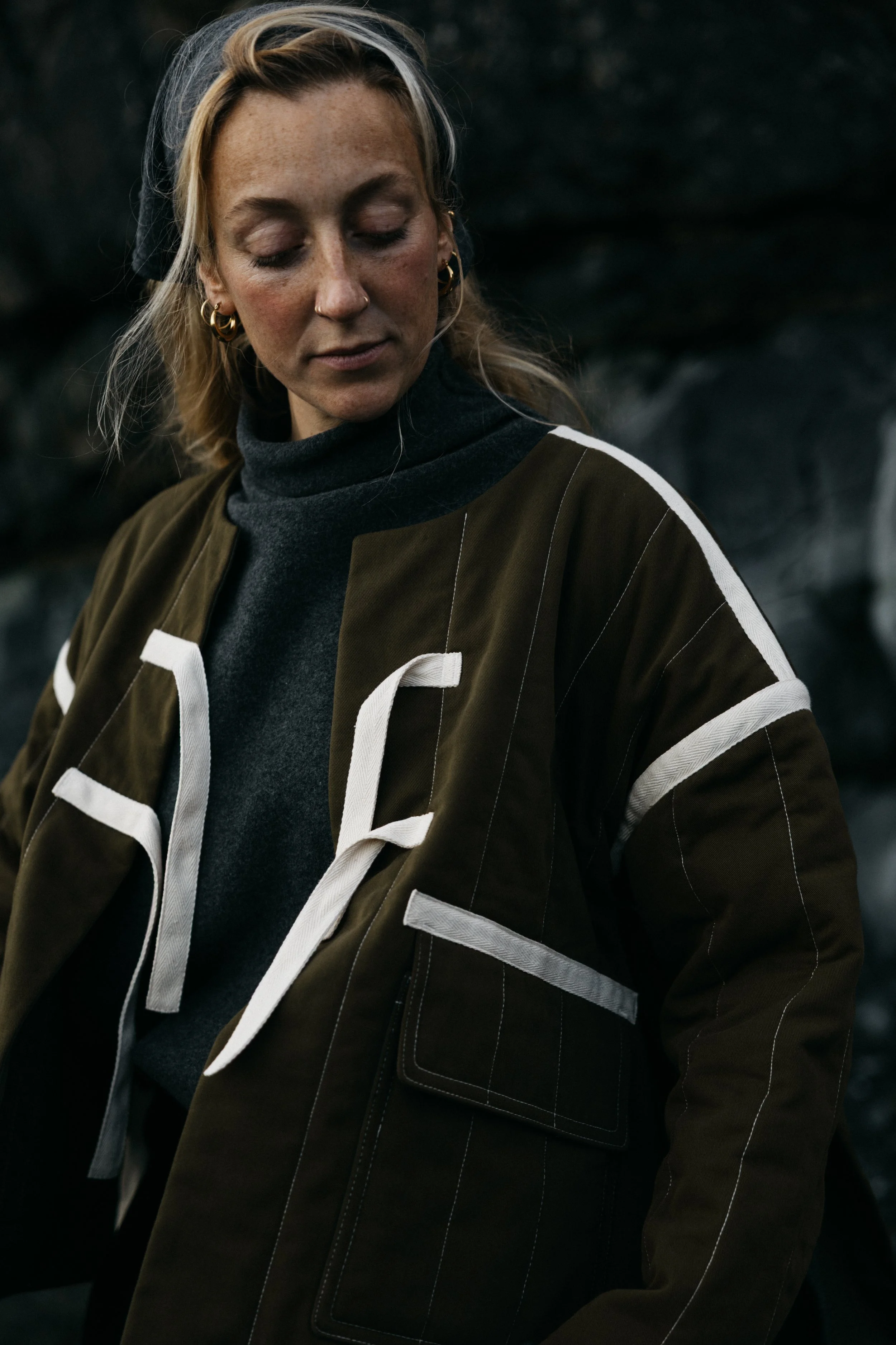 A woman with blonde hair and light freckles, wearing a dark green jacket with white accents and a dark gray turtleneck shirt, standing outdoors against a dark, rocky background, looking down with a contemplative expression.