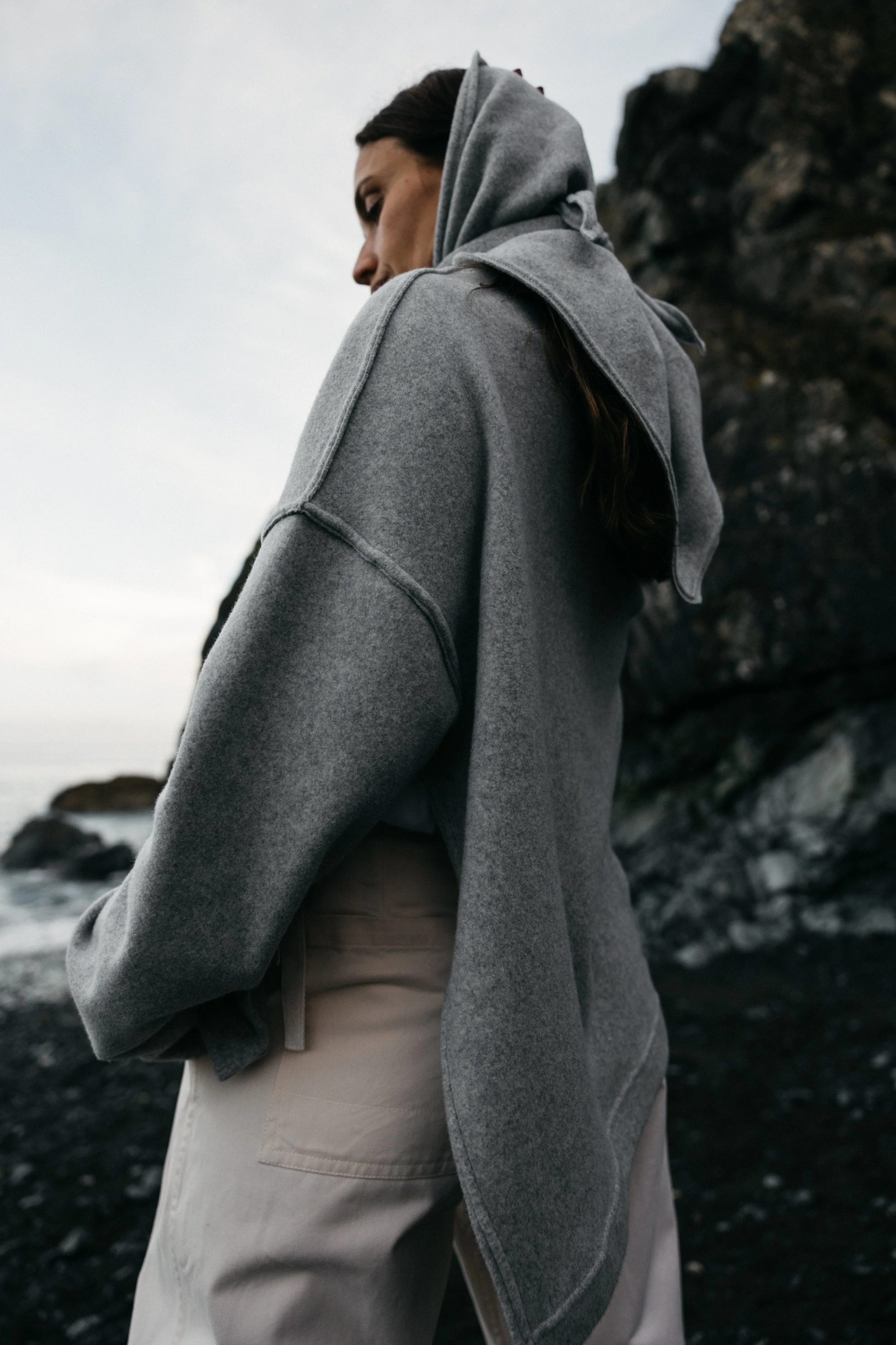 A woman in a gray hoodie and beige pants walking along a rocky shoreline with dark cliffs and an overcast sky.