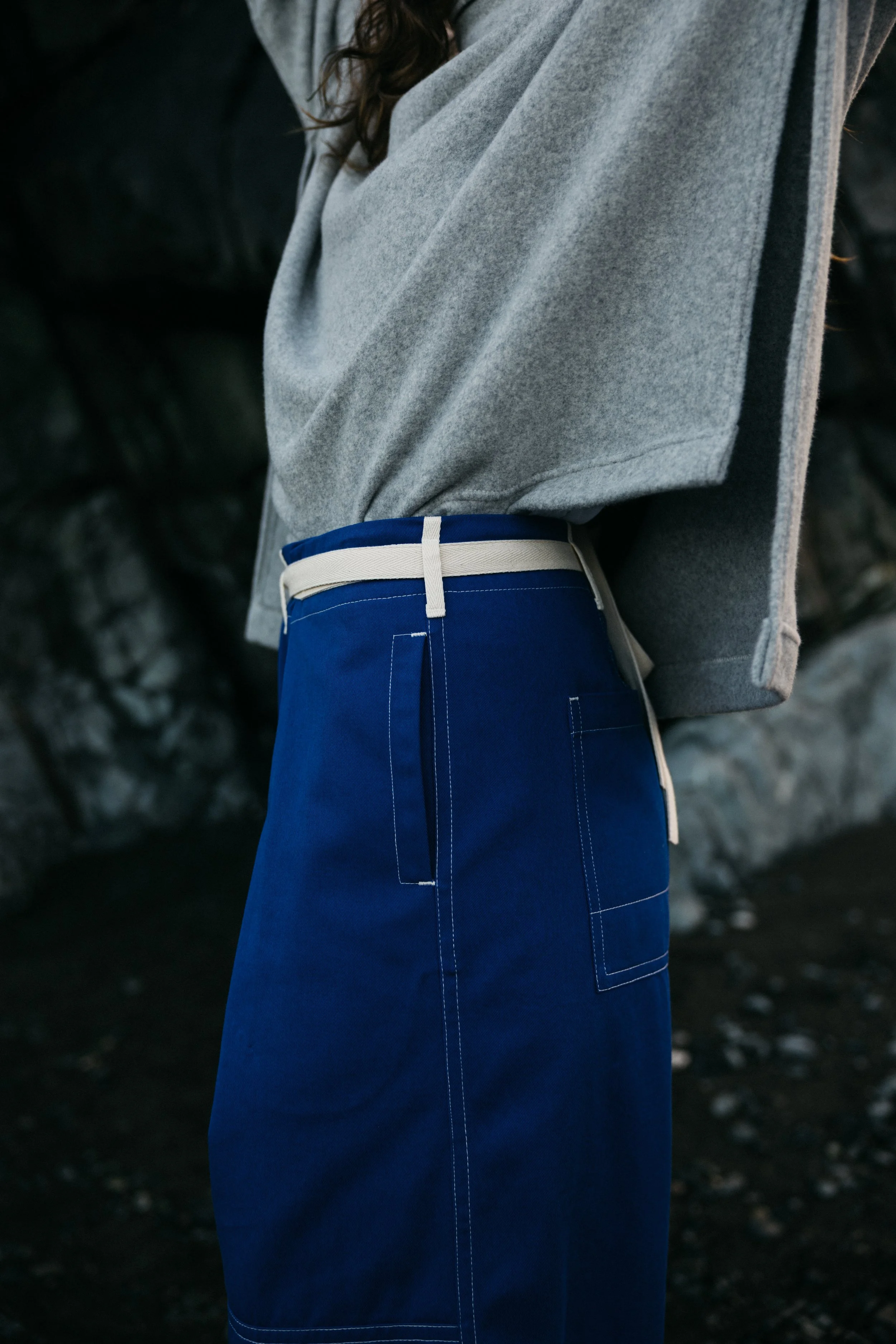 Close-up of a person wearing a gray sweatshirt and blue pants with white stitching and a beige belt, standing near a rocky background.