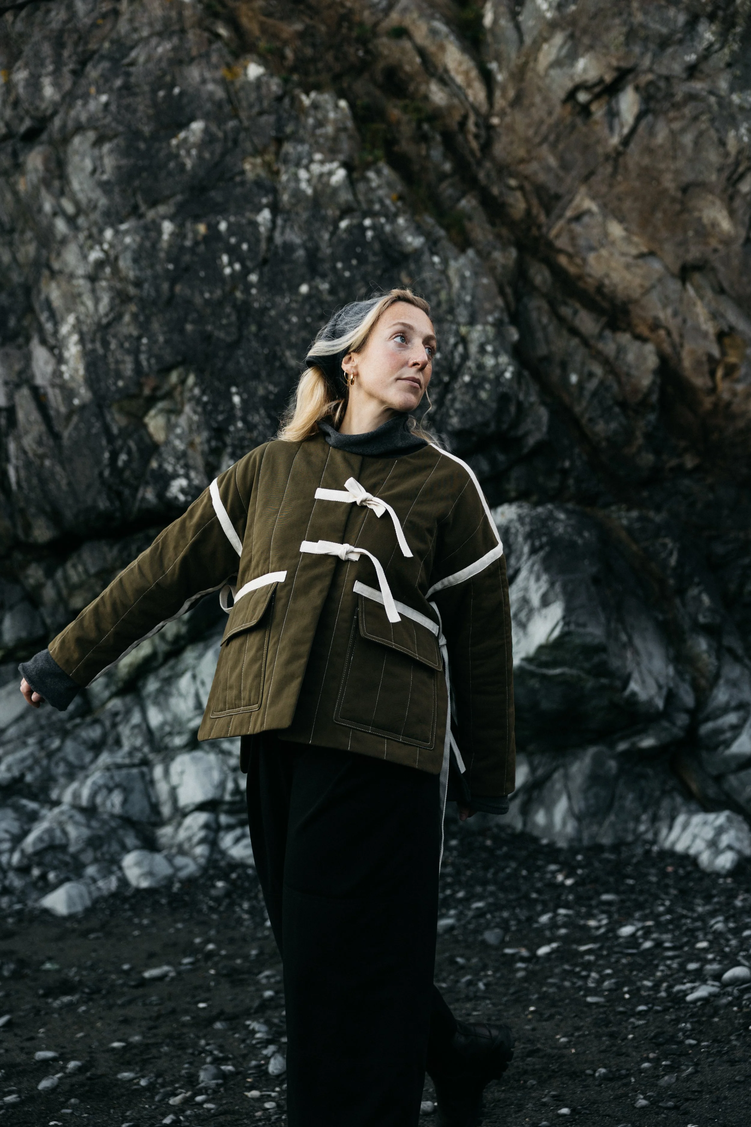 A woman standing outdoors near a rocky cliff face, wearing a brown coat with white accents and black pants, looking to the side.