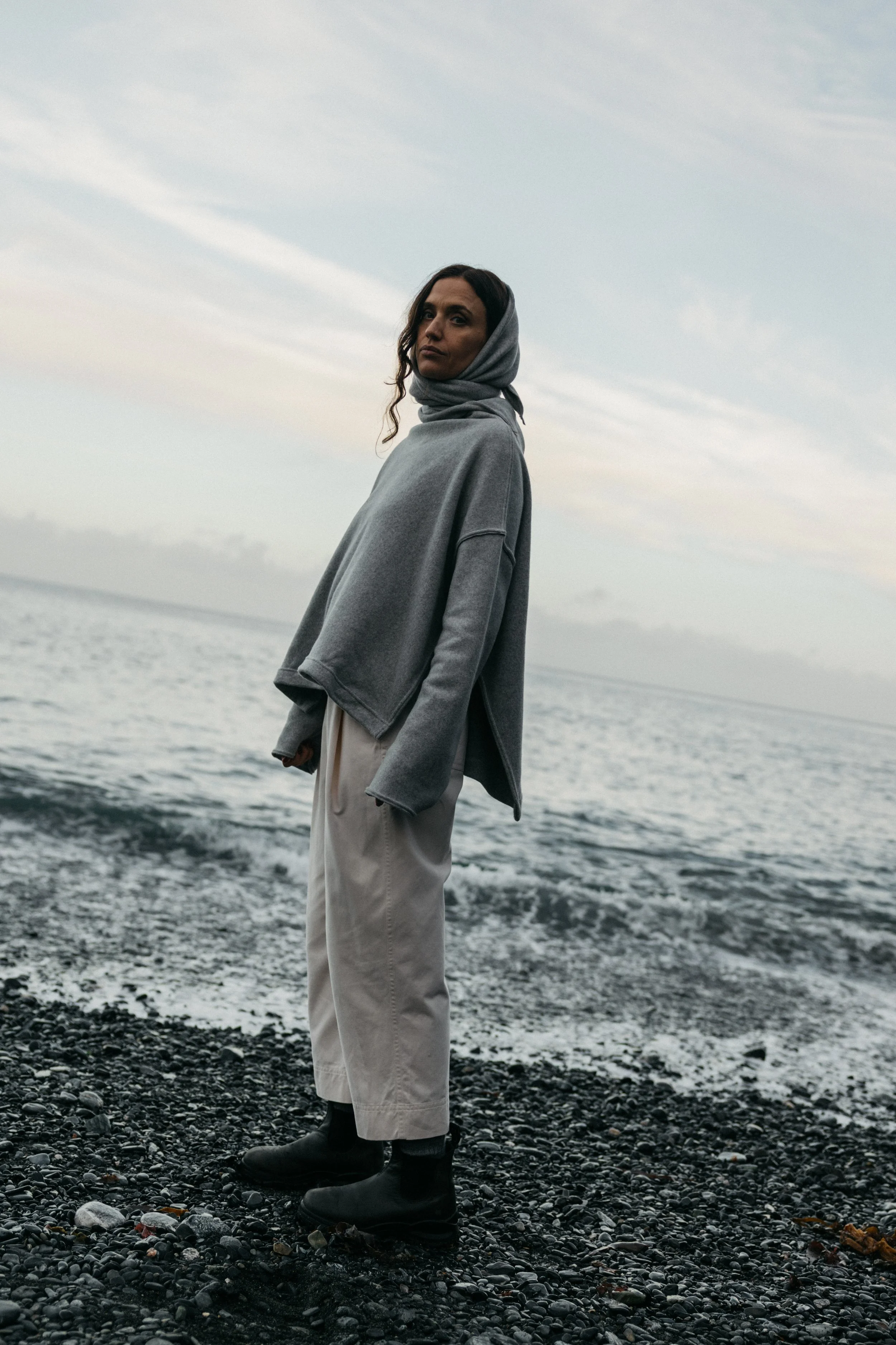 Woman standing on a rocky beach near the water's edge, wearing a gray hoodie, beige pants, and black boots, with cloudy sky in the background.