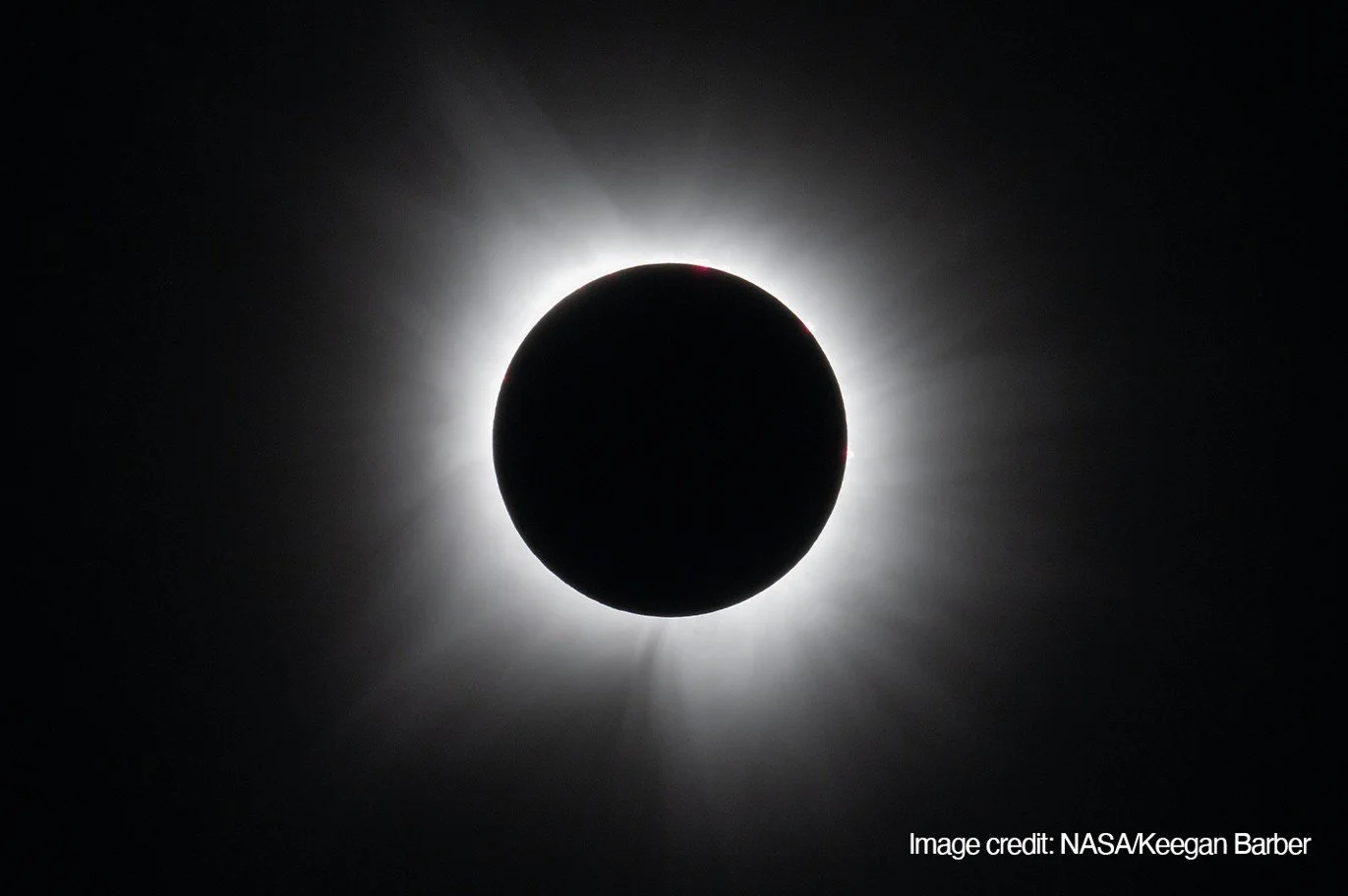 The Moon blots out the Sun during a total eclipse, revealing the corona as a pale, uneven halo of fine streamers reaching into the black sky.