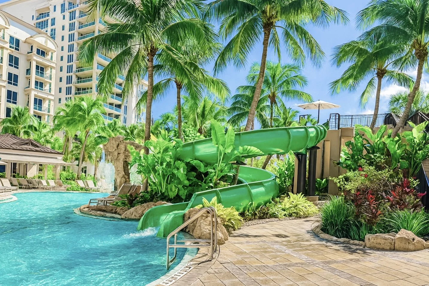 A tropical swimming pool area with a large rock structure with a waterfall, green water slide, palm trees, and poolside seating under a bright sky.