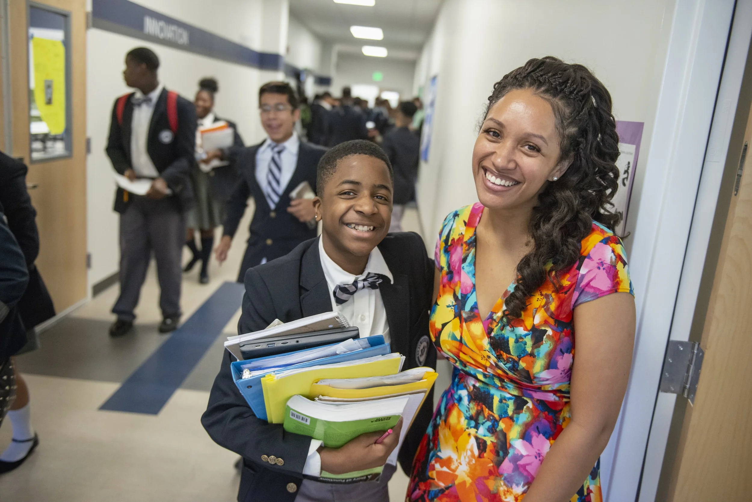A smiling woman and a young boy in school uniform with notebooks, standing in a hallway filled with other students.