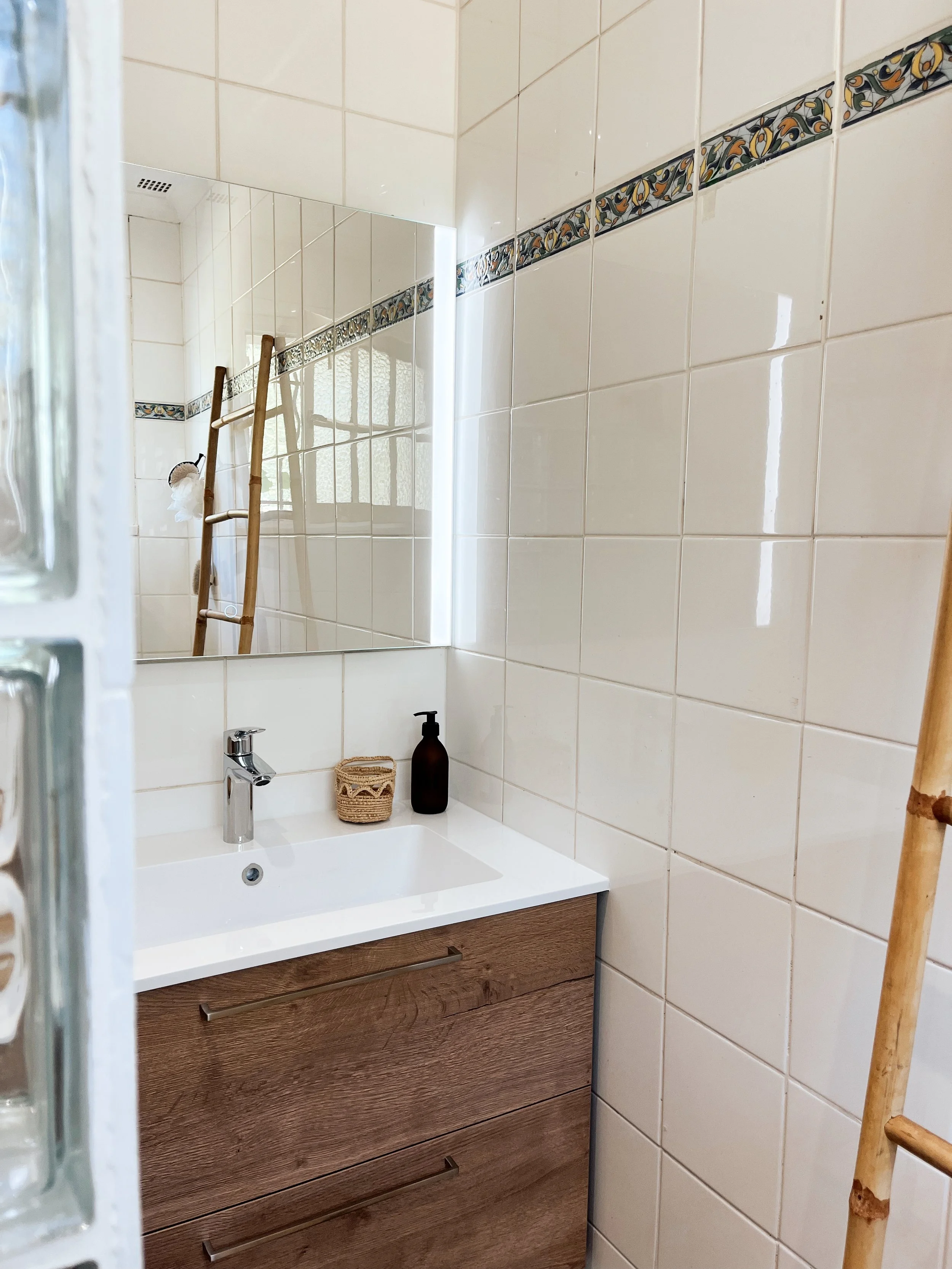 Petite salle de bain avec un miroir, un lavabo avec un meuble en bois, deux bouteilles et un porte-savon en rotin. Murs en carrelage blanc avec une bande décorative.