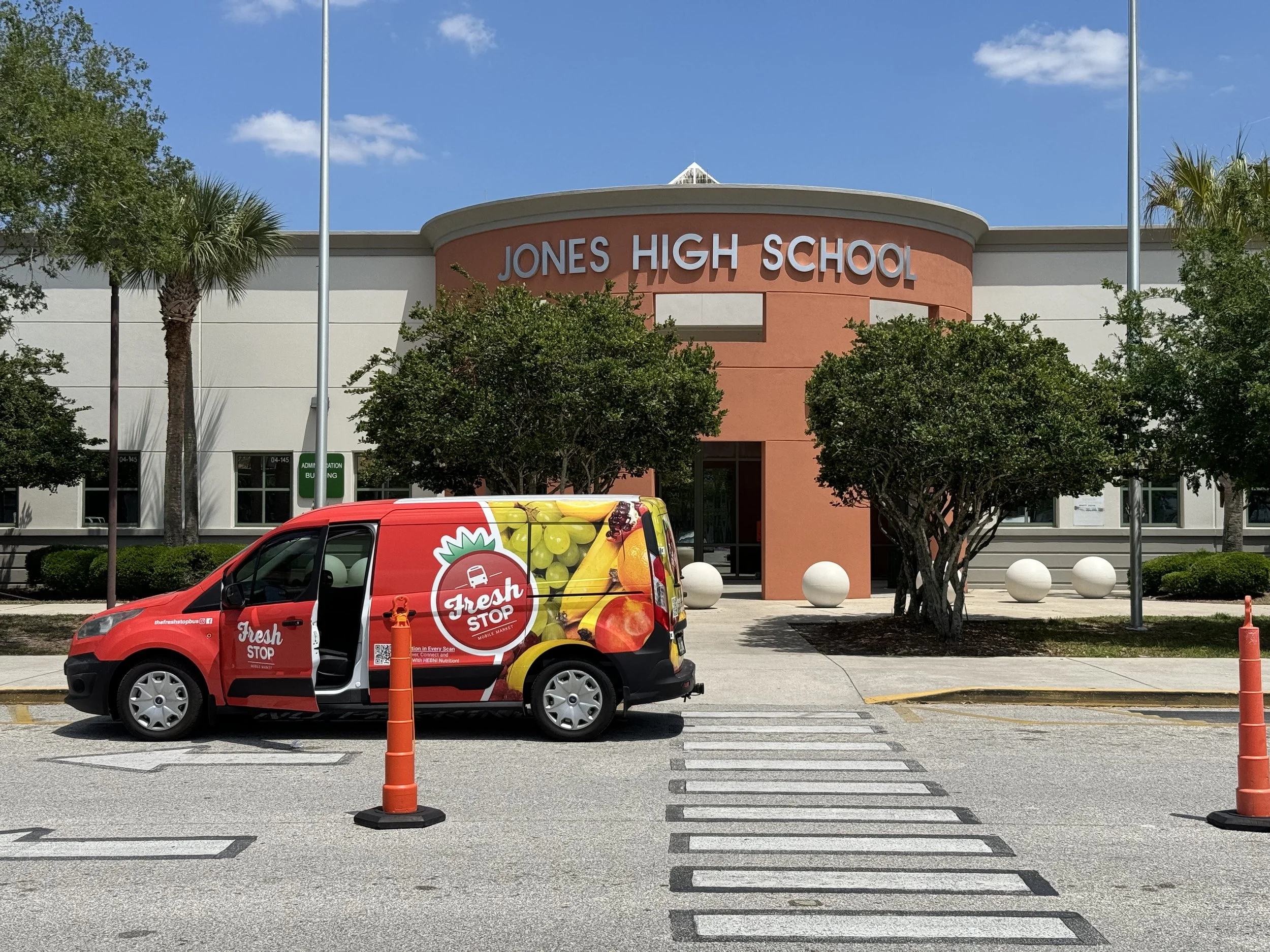 Jones High School with a colorful van in the foreground, marked as Fresh Stop, parked at a crosswalk on a sunny day with clear skies.