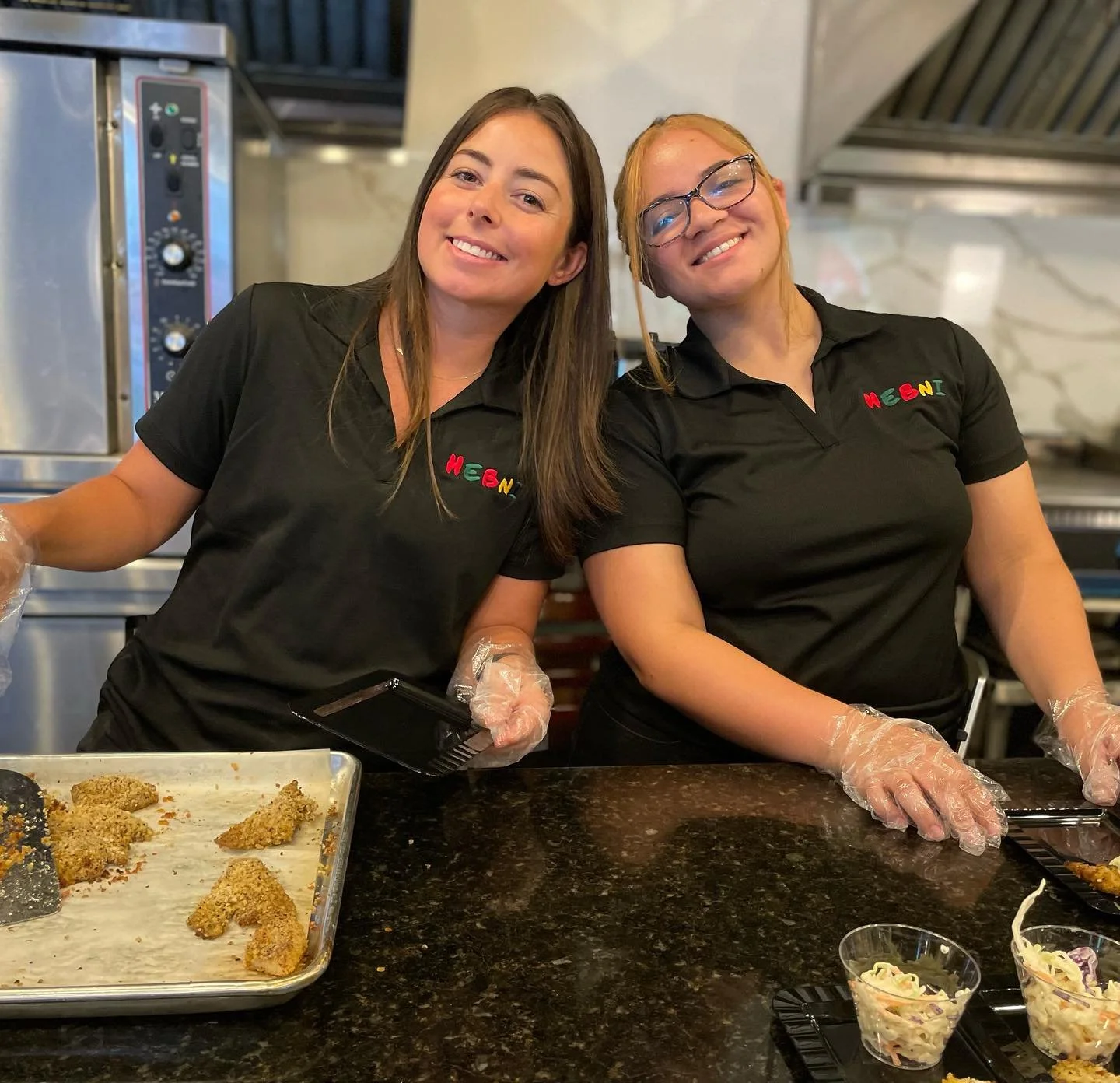 Two women in black shirts with colorful logo, wearing plastic gloves, smiling and working in a kitchen with food and cooking utensils.