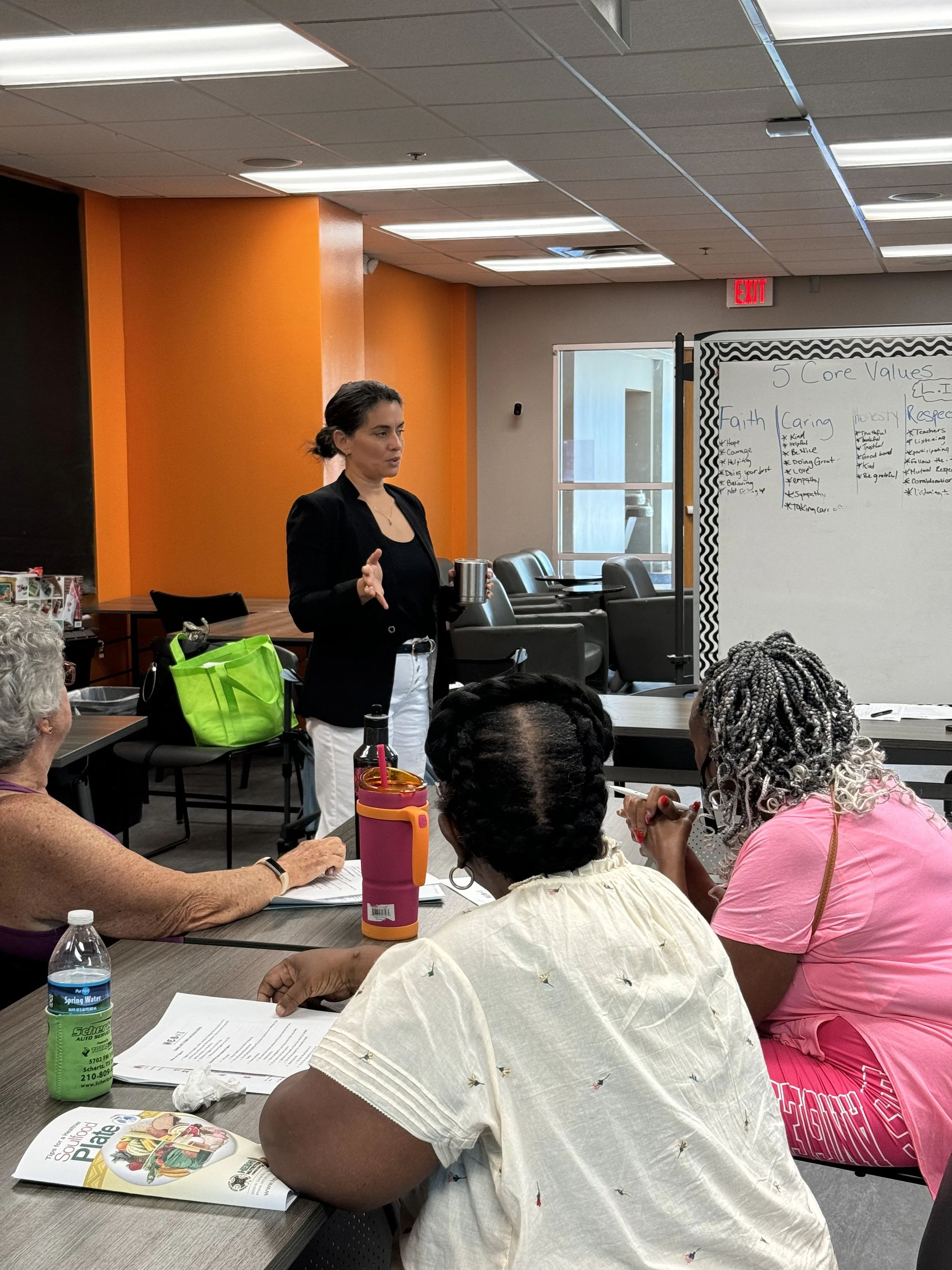 A woman in a black blazer standing and speaking in front of a small group of women sitting at a table during a meeting or presentation in a room with orange and beige walls. There is a whiteboard with writing and a window in the background.