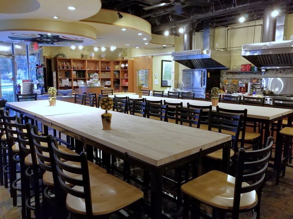 Empty restaurant with long communal tables decorated with small flower arrangements, kitchen area with stainless steel appliances and hanging range hoods, and bookshelves in the background.