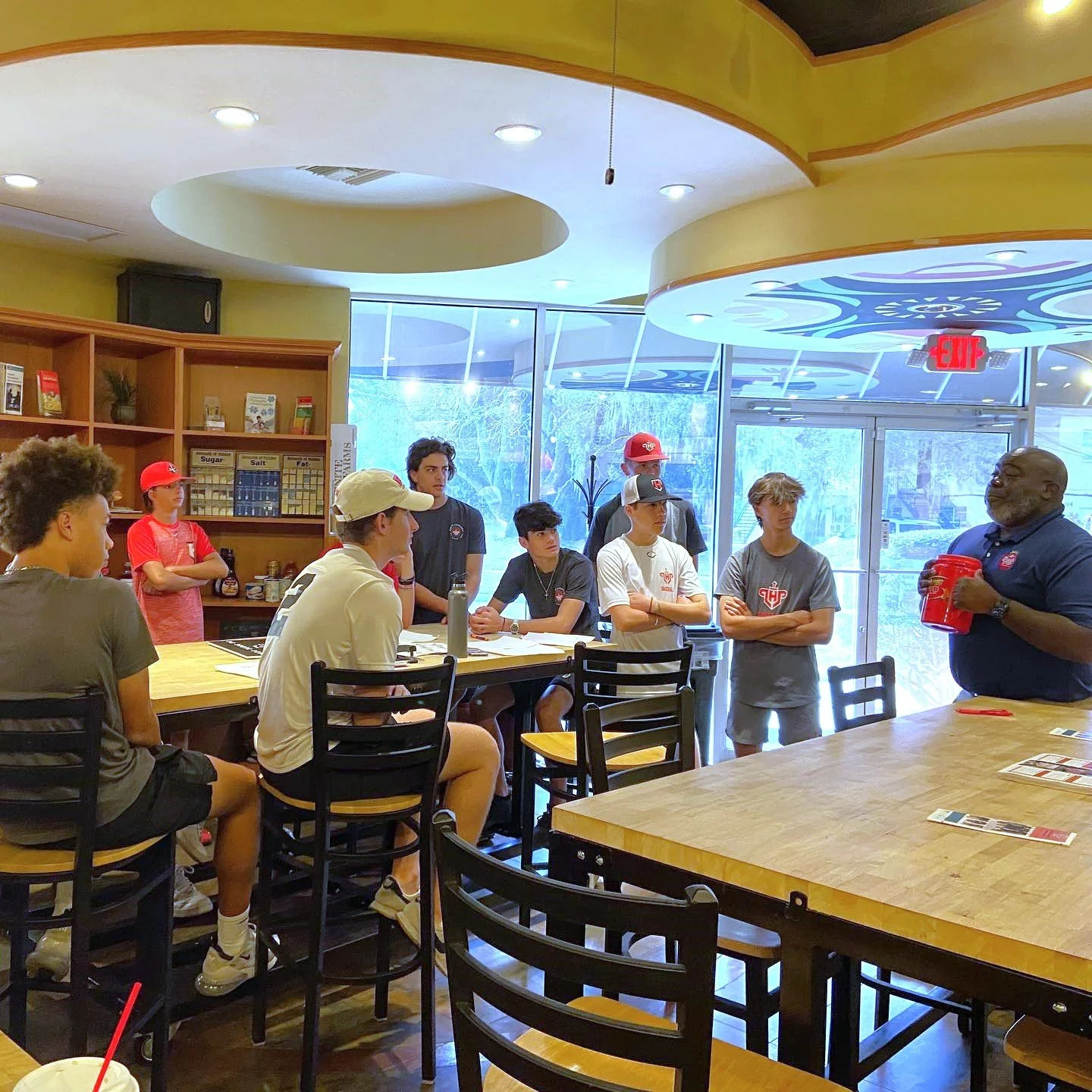 Group of young people sitting and standing around a counter in a brightly lit restaurant or cafe, with a man speaking to them holding a red container, and shelves with spices and food items in the background.
