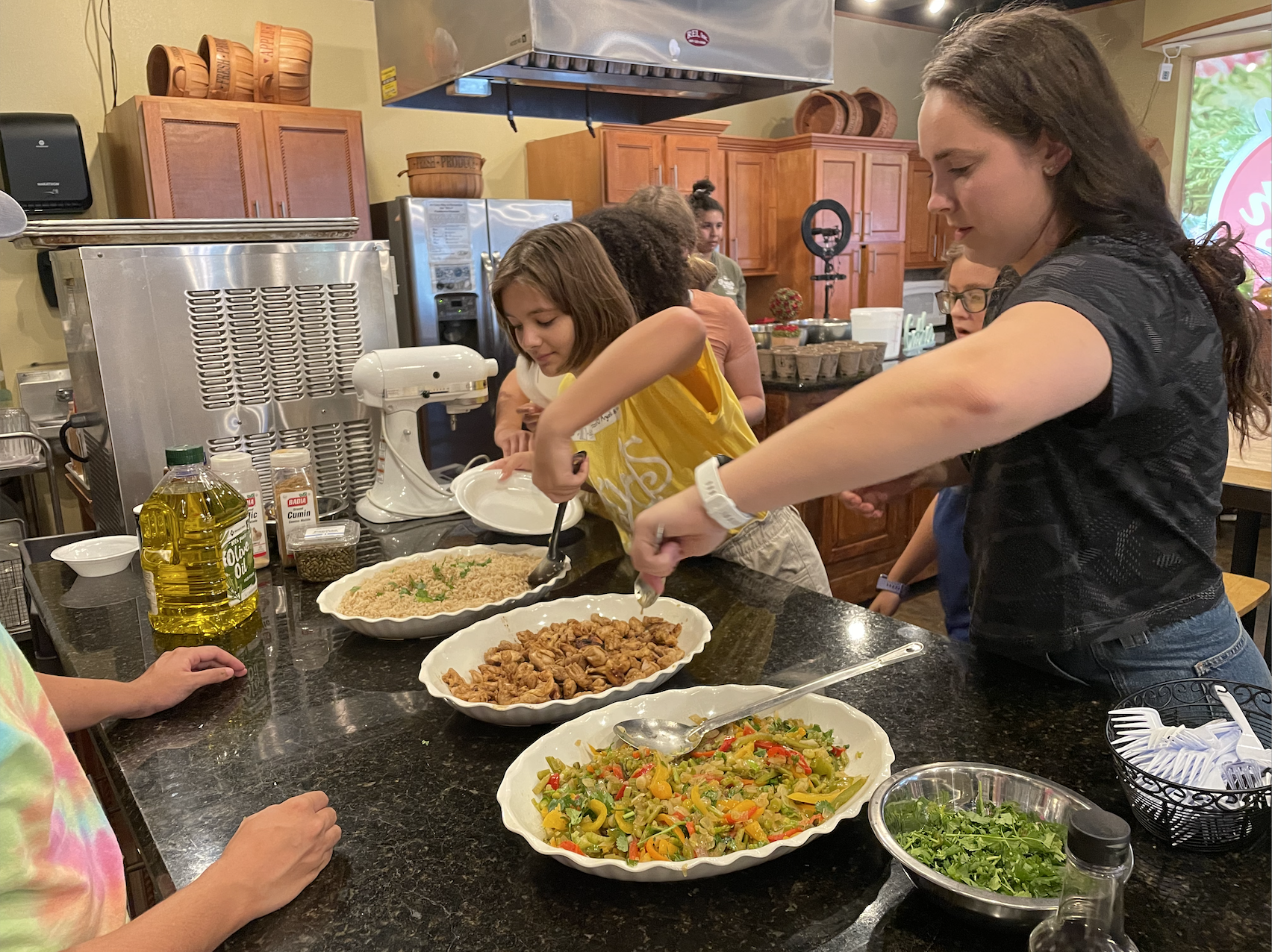 People serving and preparing various dishes in a kitchen or dining area, including salads, cooked chicken, and rice.