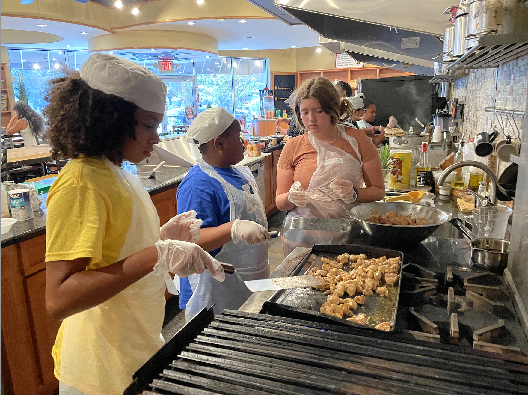 A group of young girls wearing aprons and caps cooking chicken on a grill inside a kitchen at a restaurant.