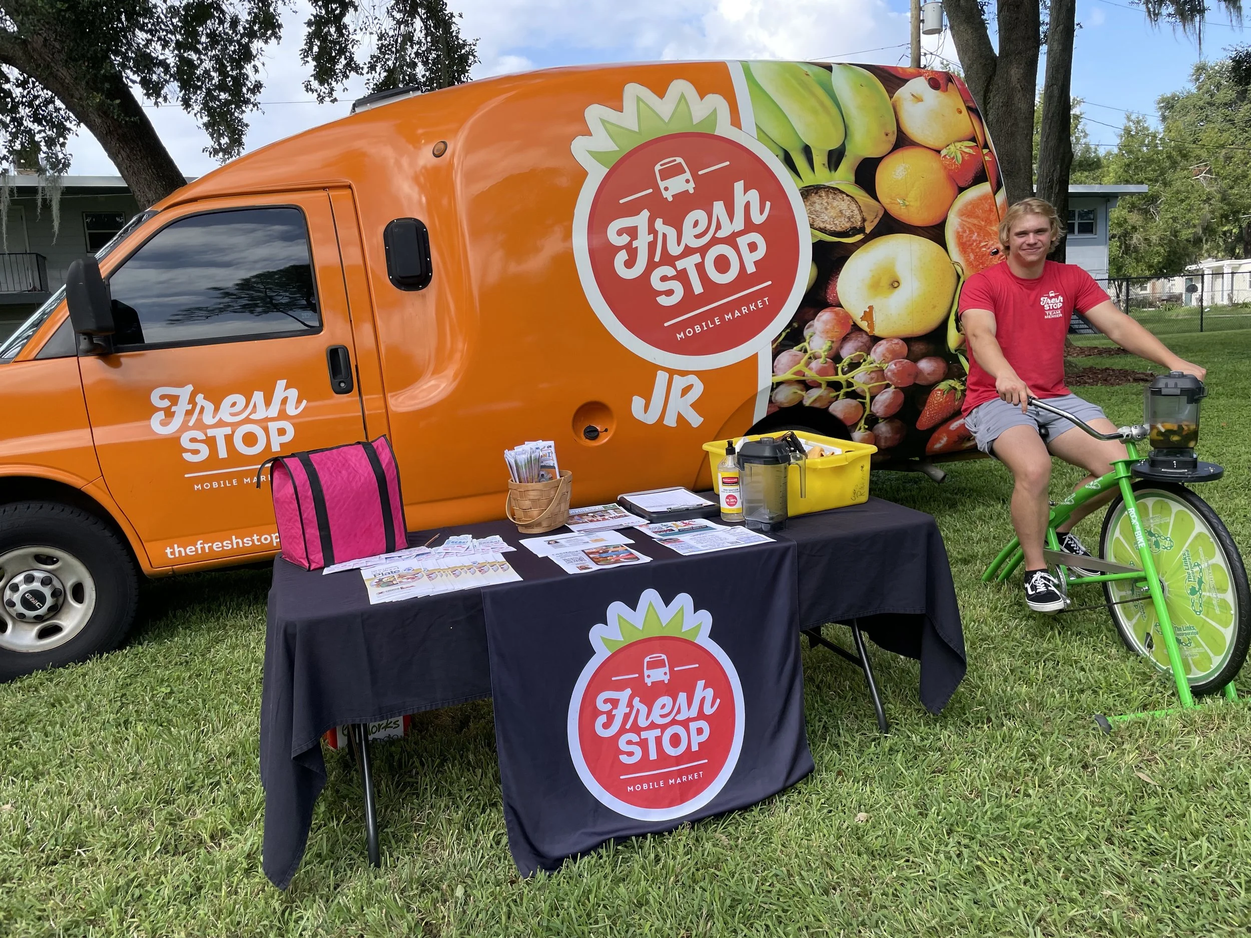 A young man on a green bicycle standing next to a mobile market truck labeled 'Fresh Stop' with a large fruit graphic on the side. The bright orange truck has the logo 'Fresh Stop' and a table in front with market materials.