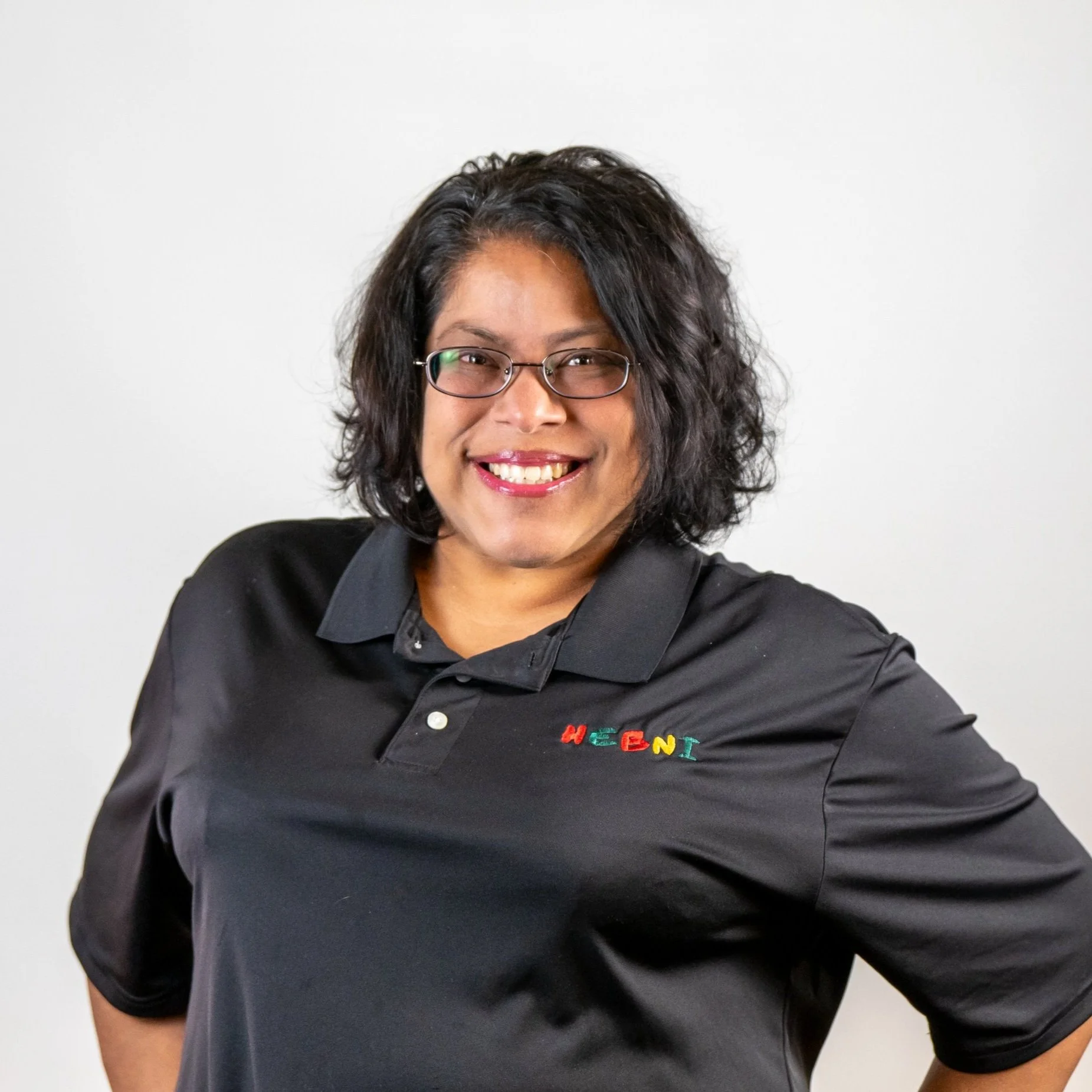 A woman with shoulder-length curly black hair, wearing glasses and a black polo shirt with colorful embroidered letters on the chest, standing against a plain white background, smiling.