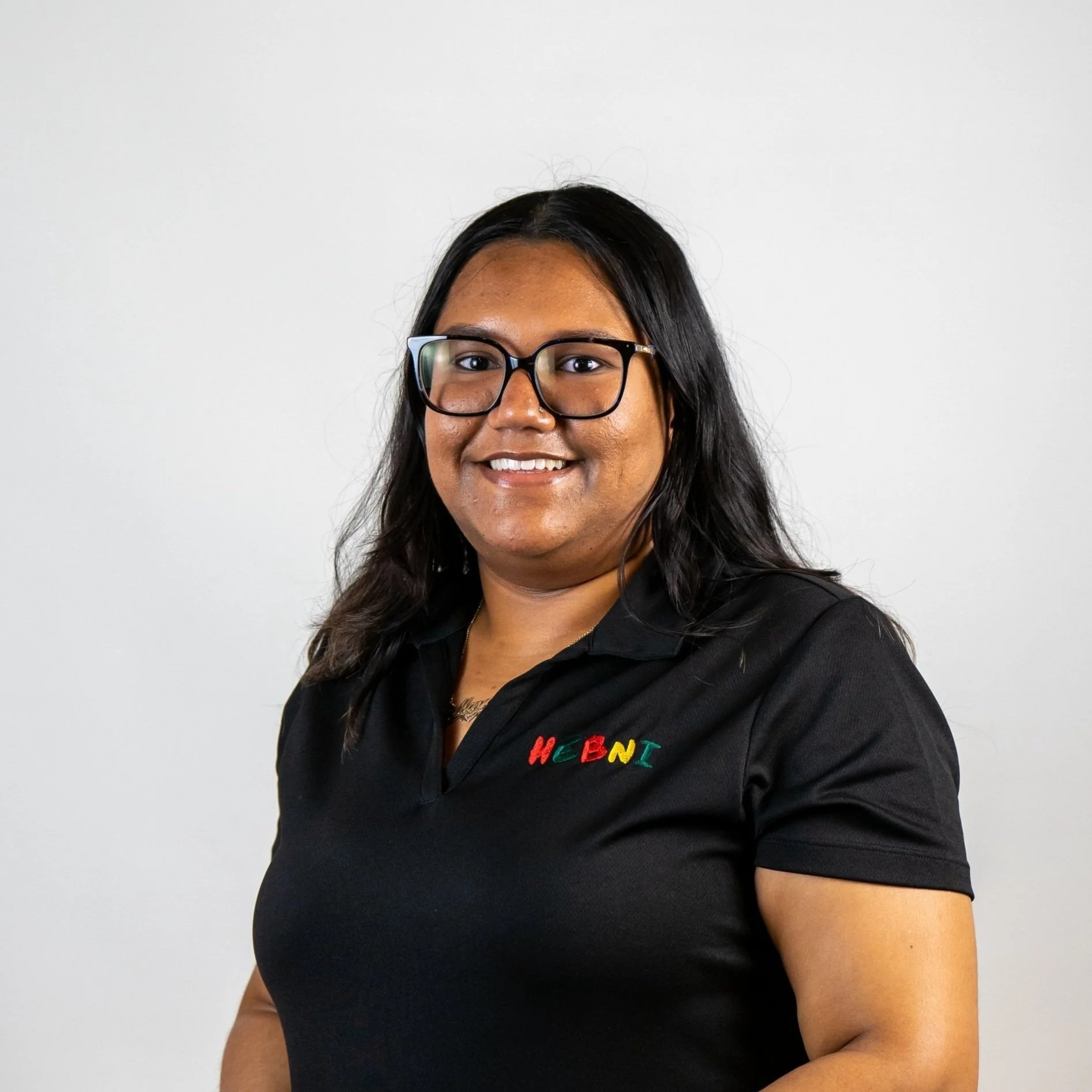 A smiling woman with long black hair, glasses, and wearing a black polo shirt with colorful embroidered lettering that spells 'WEBNI'.