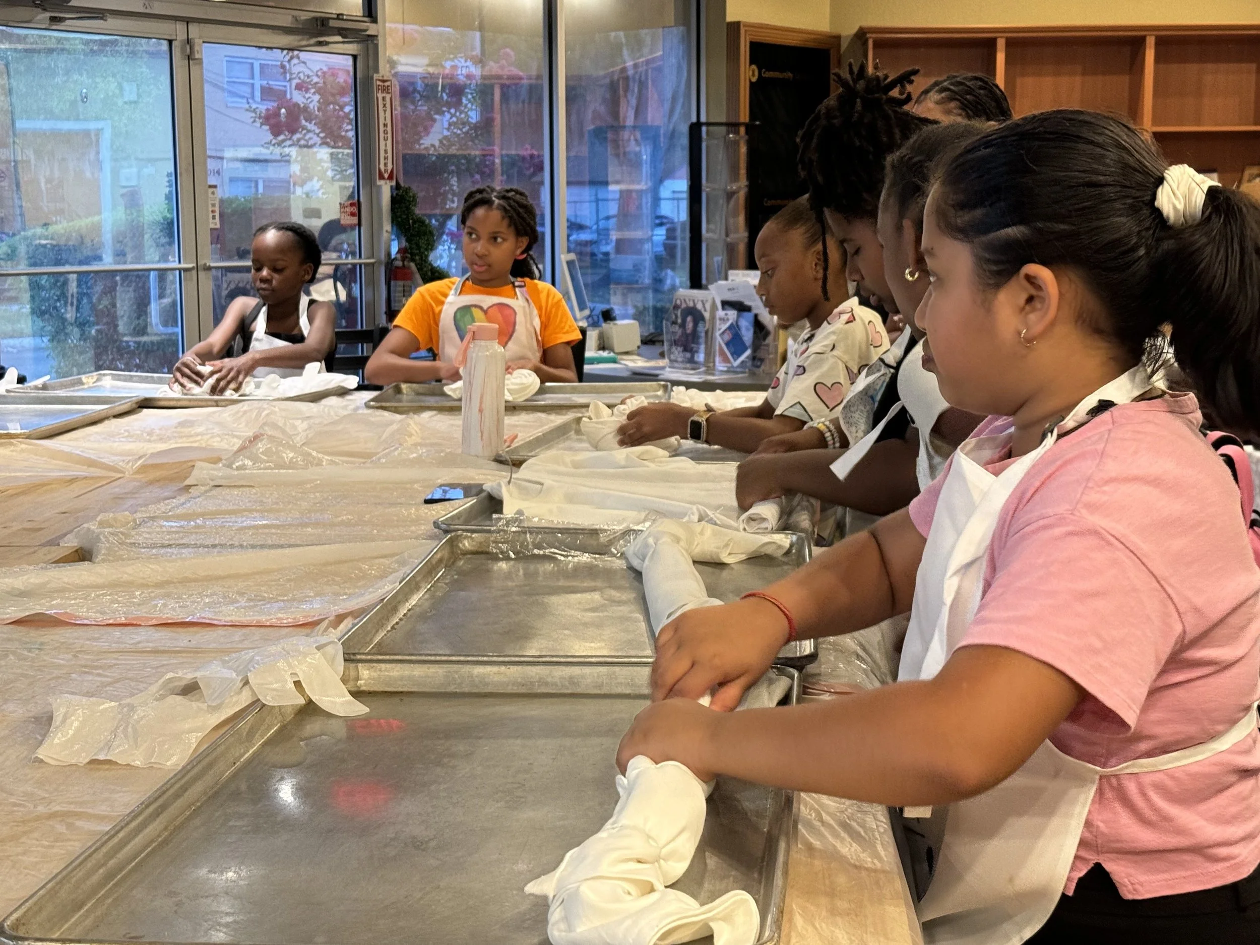 Children participating in a cooking class, wearing aprons and gloves, gathered around a table with baking trays and cooking supplies in a kitchen or classroom setting with large windows and wooden shelves.