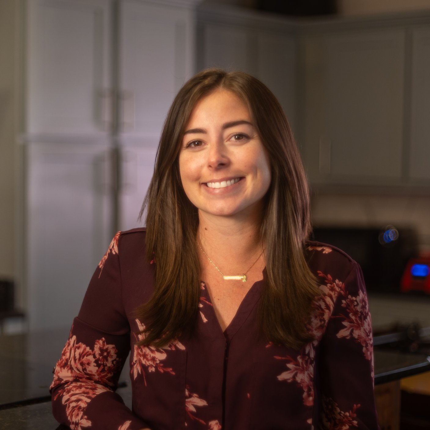 A smiling woman with brown hair in a kitchen, wearing a burgundy floral blouse and a gold necklace.