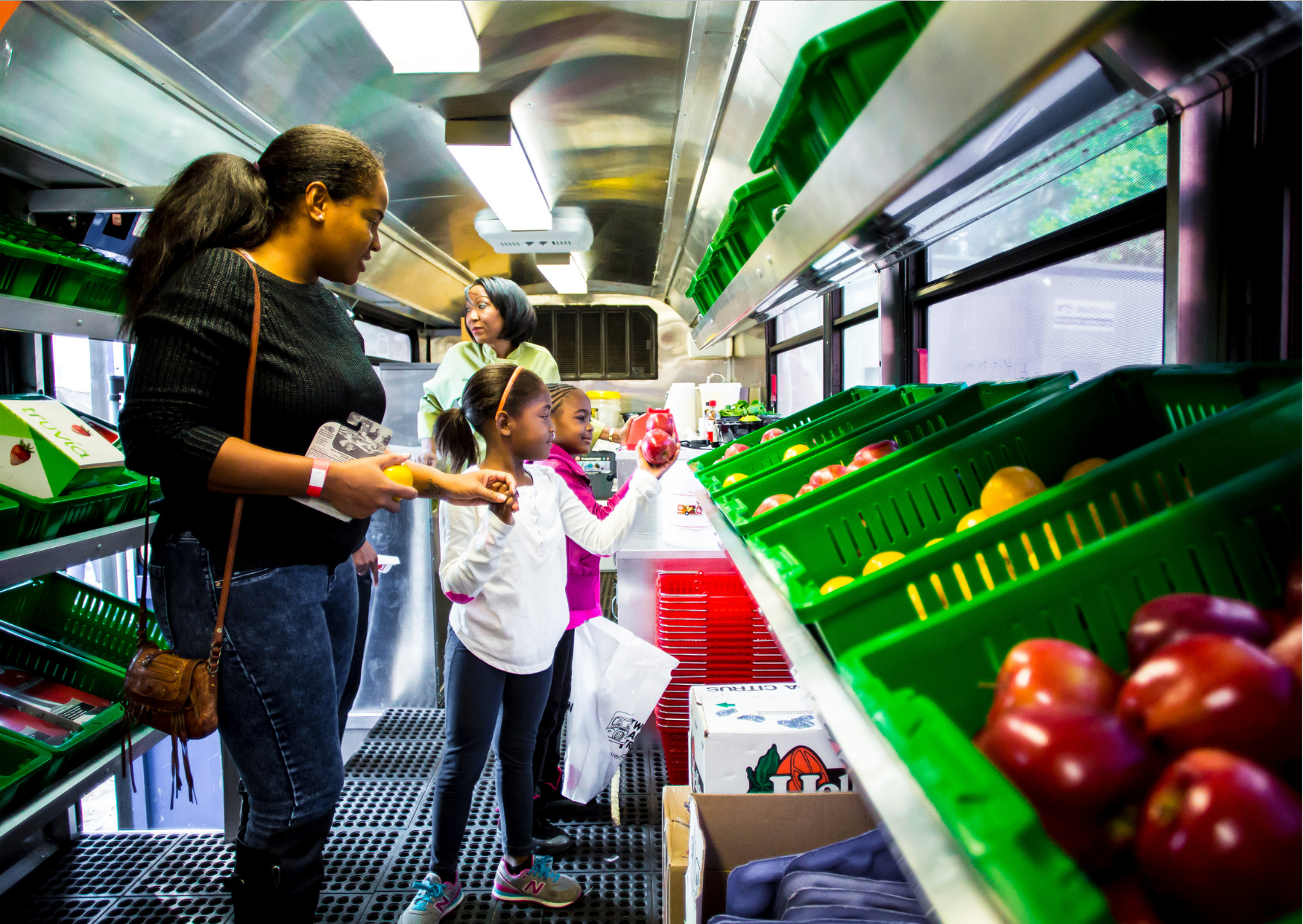 A group of people, including children and adults, shopping for fruits and vegetables inside a mobile market. They are examining produce such as tomatoes and apples in green baskets on shelves.