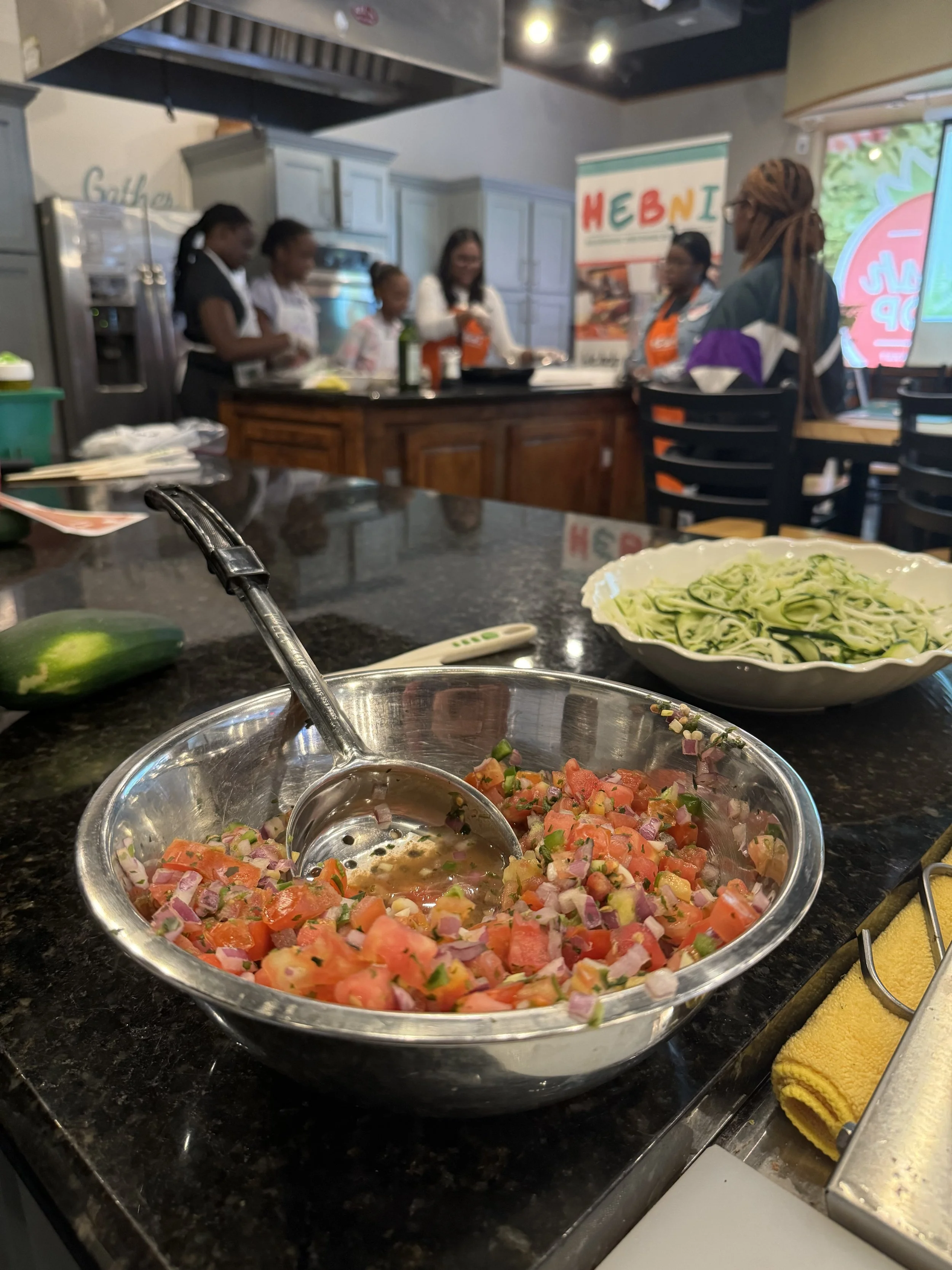 A stainless steel bowl filled with chopped tomatoes, onions, and green peppers, with a serving spoon inside. In the background, there is a bowl of spiralized zucchini and a yellow towel, on a black countertop in a kitchen or cooking class setting with several people gathered around.