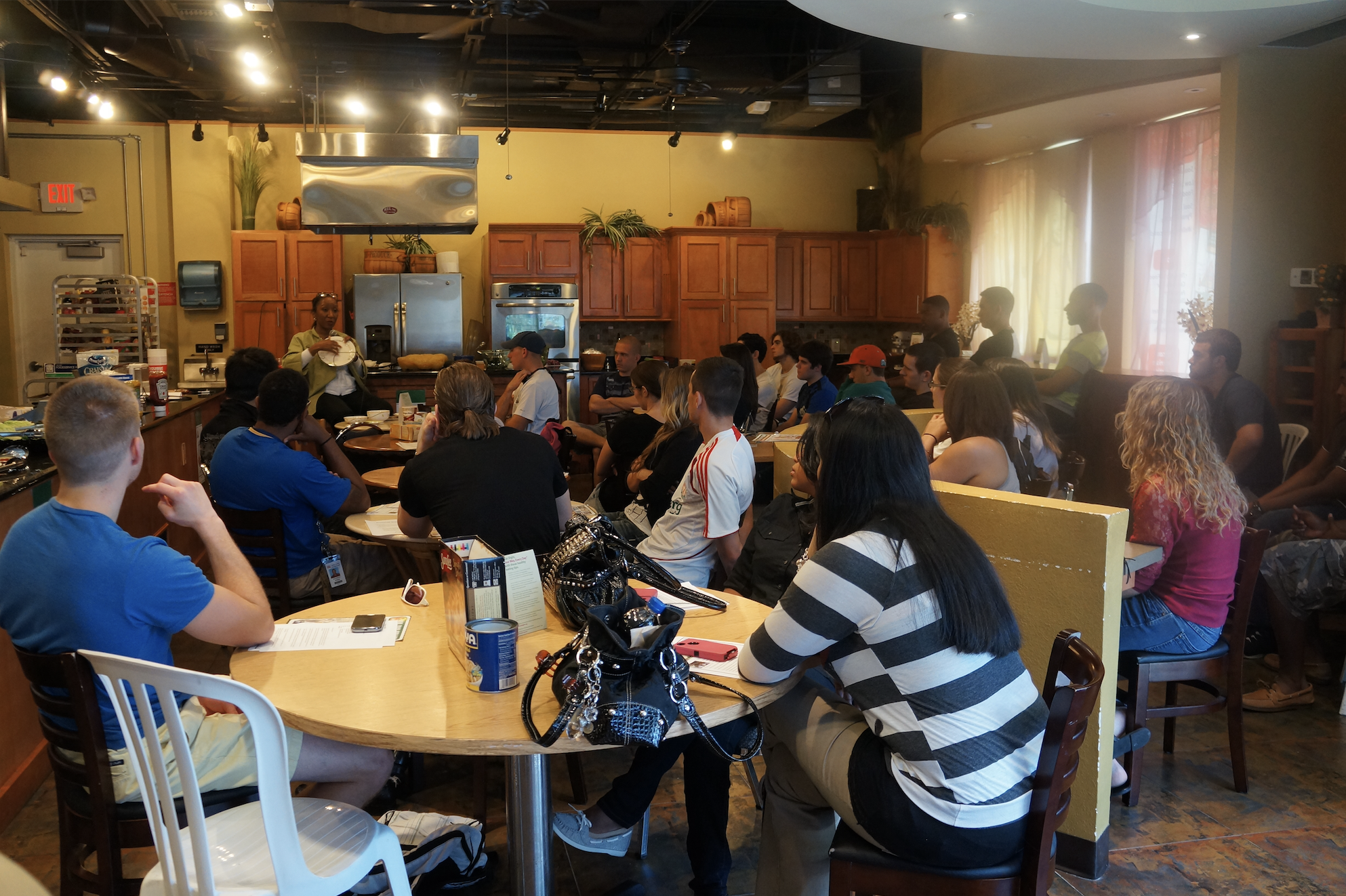 A group of people seated around tables in a kitchen-style restaurant, listening to a woman who is standing and holding a plate. The room has wooden cabinets, a stainless steel stove, and is illuminated with warm lighting.