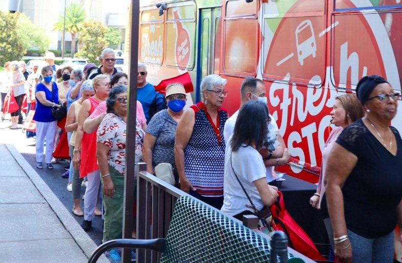People standing in line outdoors next to a colorful van with 'Fresh' written on it, some wearing masks and casual clothes.