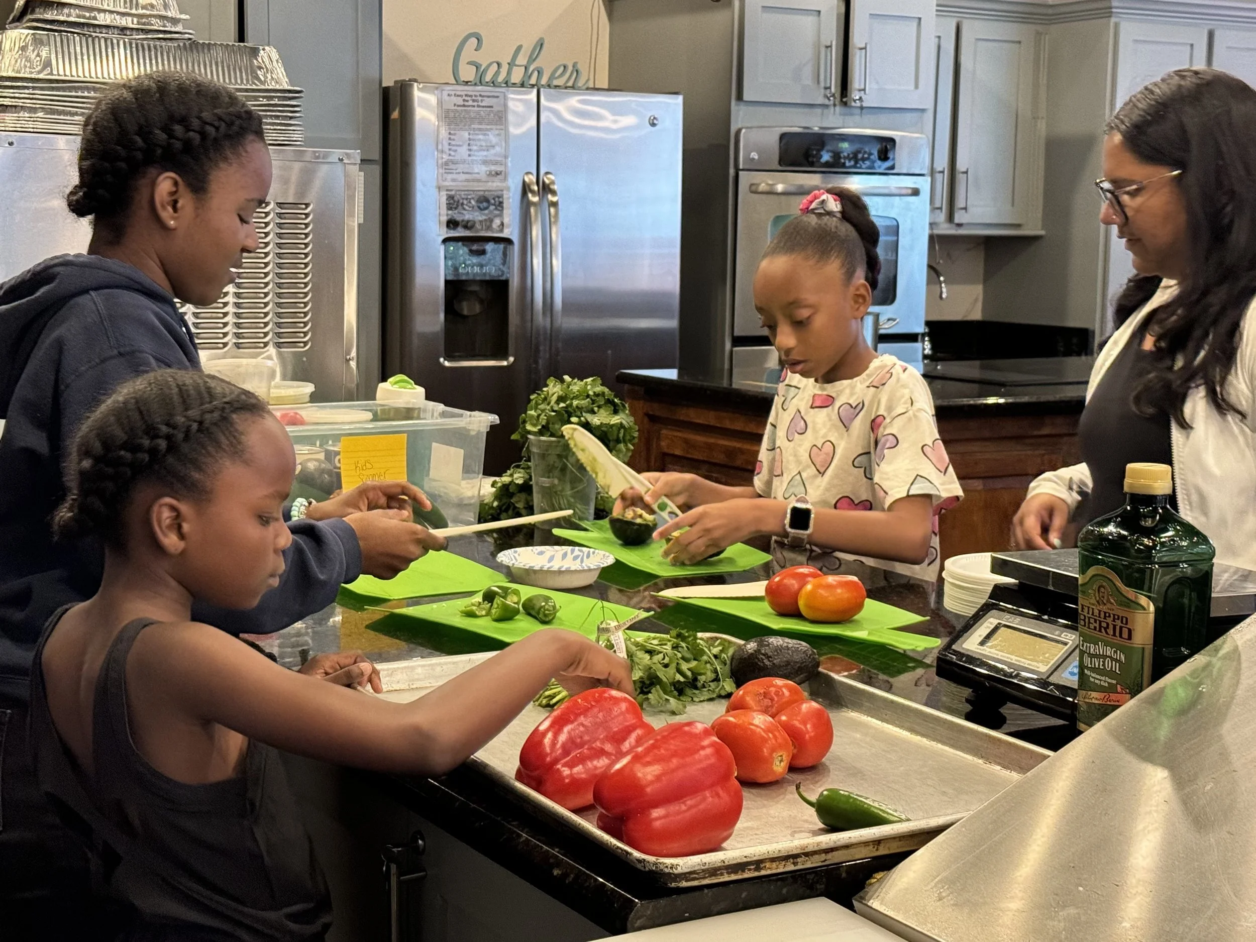 A group of five people, including children and an adult woman, are preparing vegetables in a kitchen. They are chopping and arranging tomatoes, peppers, and greens on a countertop with kitchen appliances and cabinets in the background.