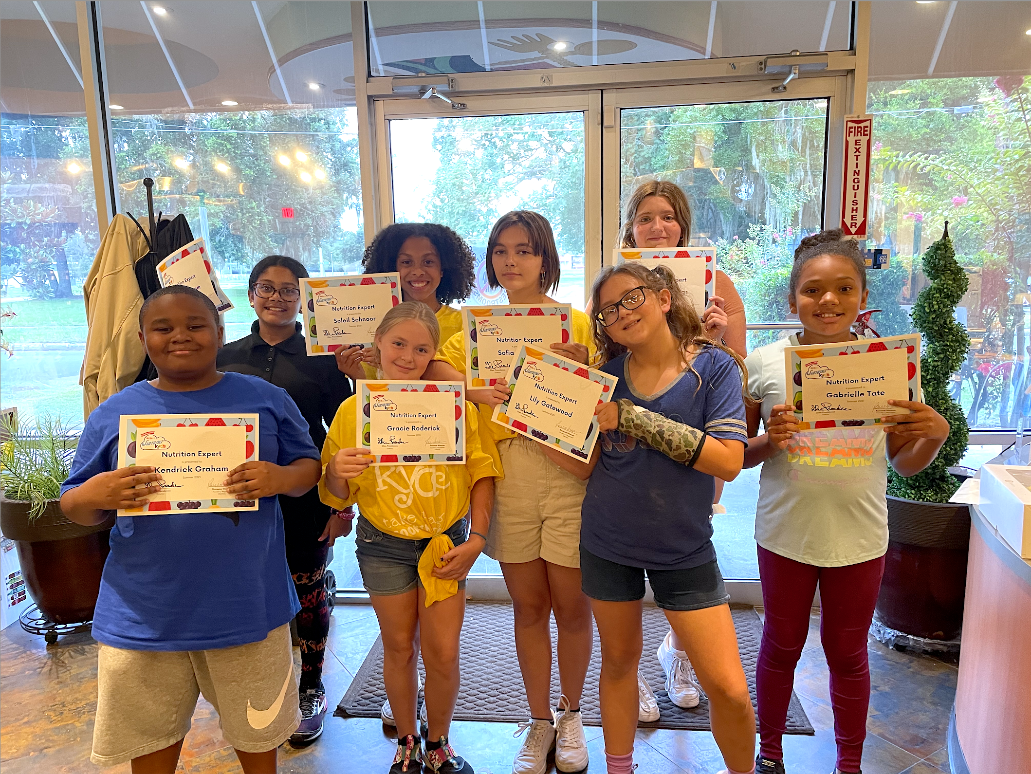 Group of children indoors holding certificates, with a glass door and plants behind them.