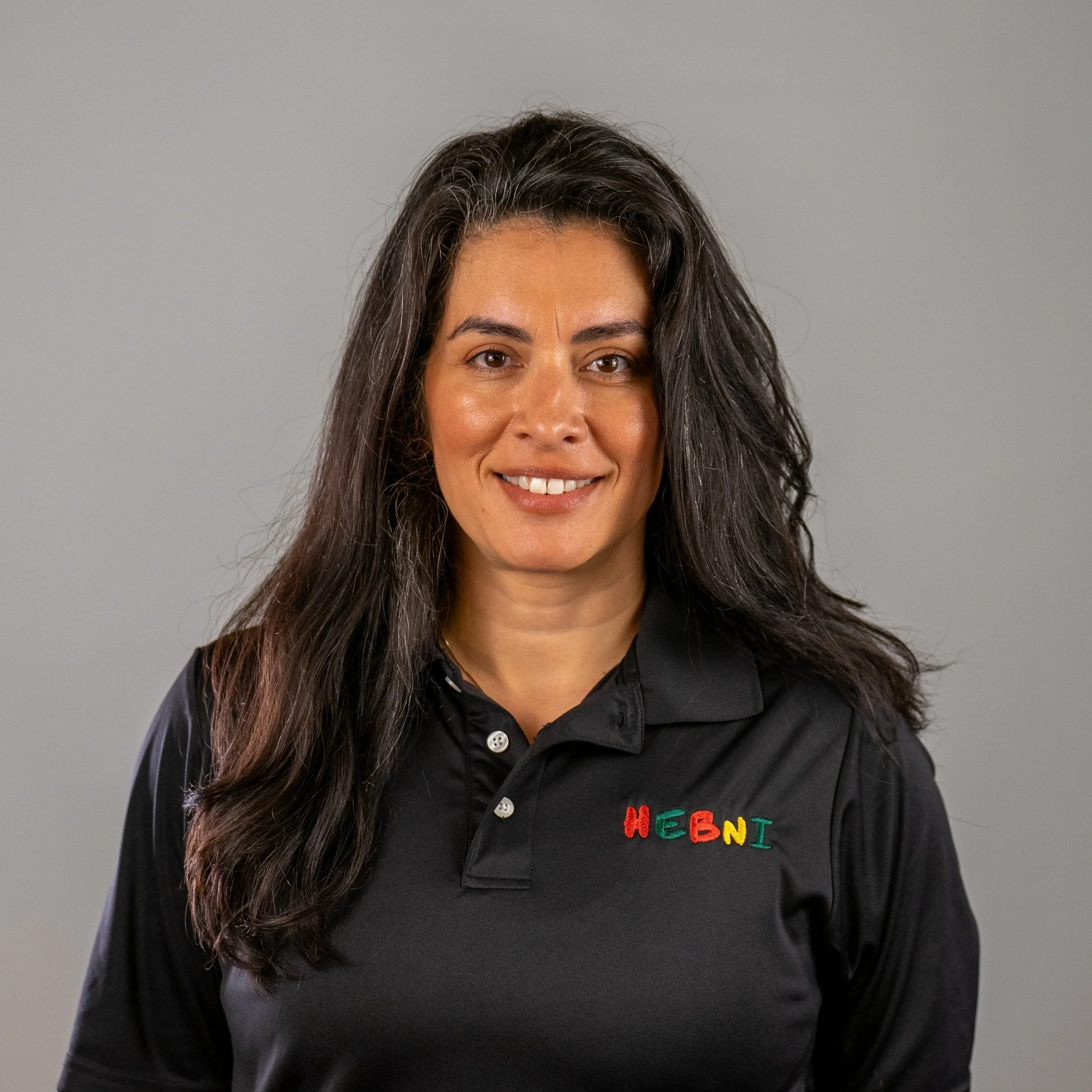 A woman with long, dark, wavy hair smiling, wearing a black polo shirt with 'HEBNI' embroidered in colorful letters on the chest, standing against a neutral gray background.