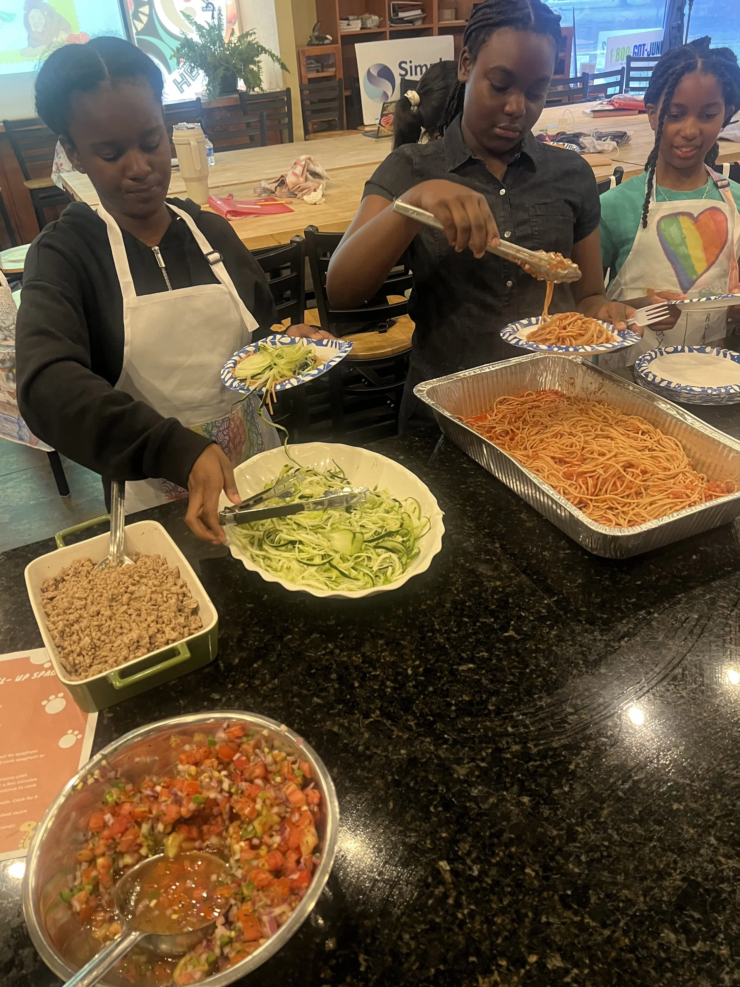 Three women in aprons serve spaghetti and salad at a buffet table in a restaurant.
