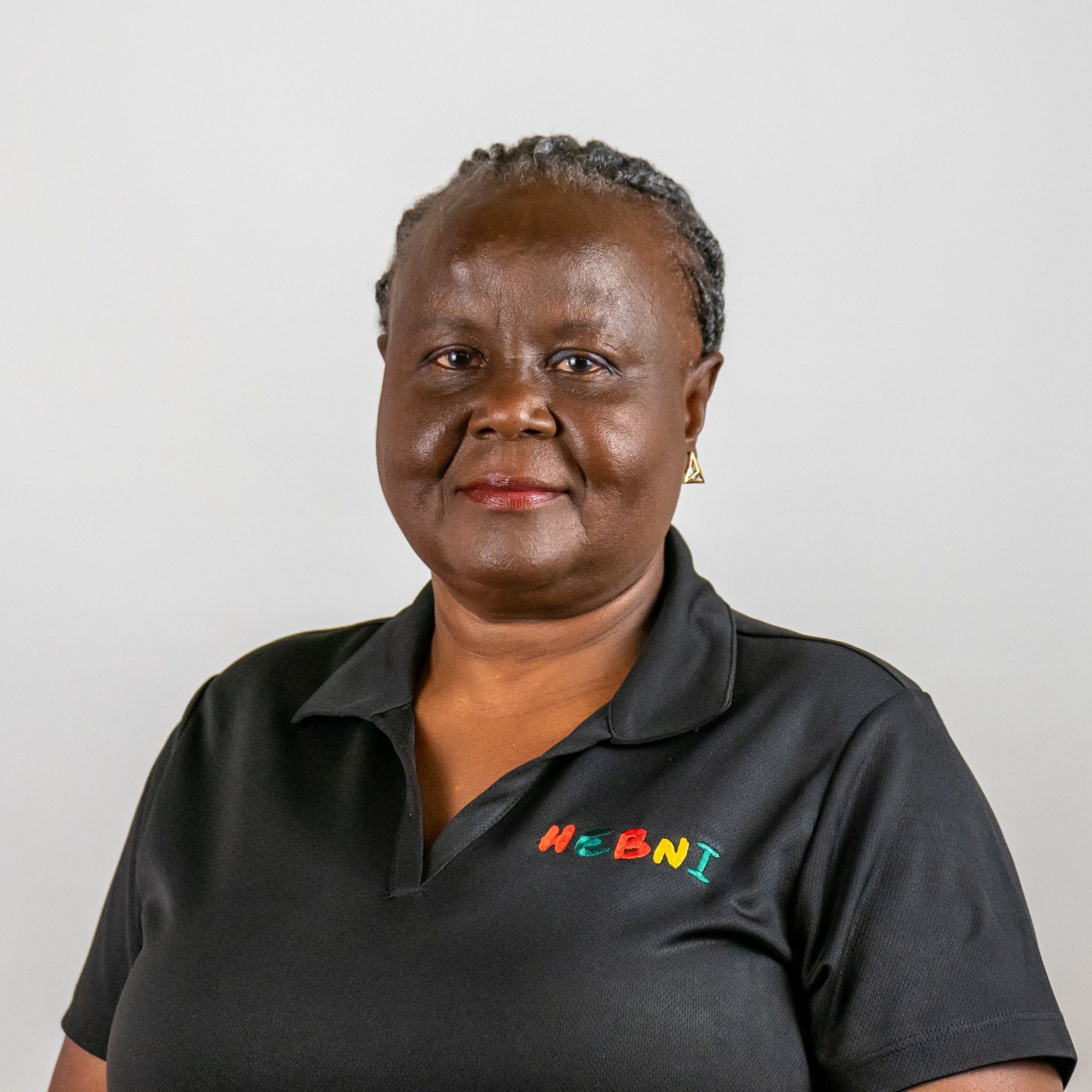 Portrait of a woman with dark skin and short hair, wearing a black polo shirt with colorful embroidery that reads 'HEBNI', standing against a plain white background.