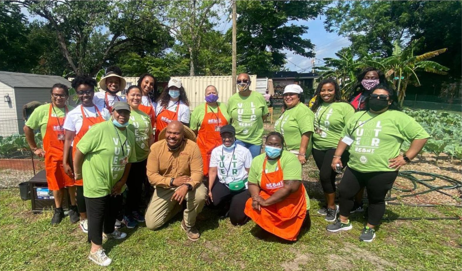 Group of diverse people in a community garden, some wearing orange aprons and others wearing green and white t-shirts with 'HEBNI' logos, posing outdoors during daytime with plants and trees in the background.