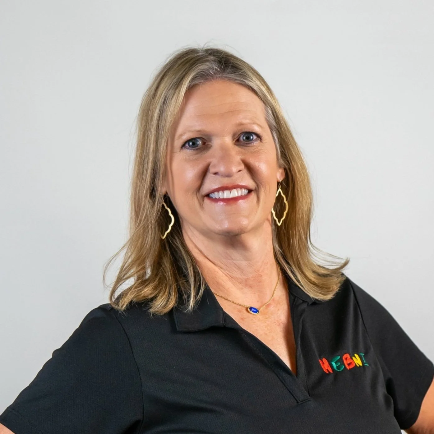 A woman with shoulder-length blonde hair smiling, wearing a black polo shirt with 'HEGN' embroidered in colorful letters, gold earrings, and a necklace with a blue pendant, standing against a plain white background.