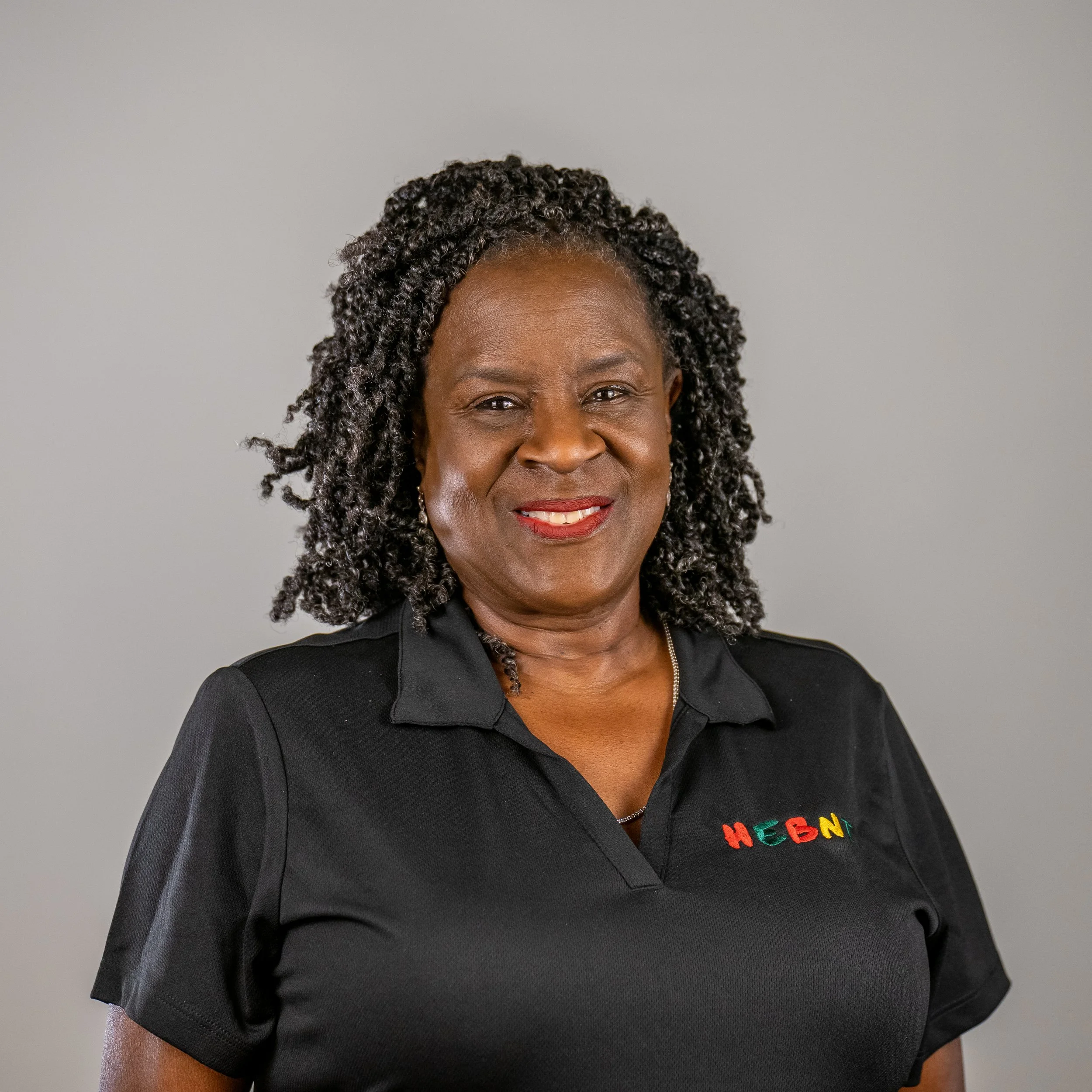 A smiling African American woman with curly black hair, wearing a black collared shirt with colorful embroidered letters that spell 'HEBNI' on the chest, standing against a plain gray background.