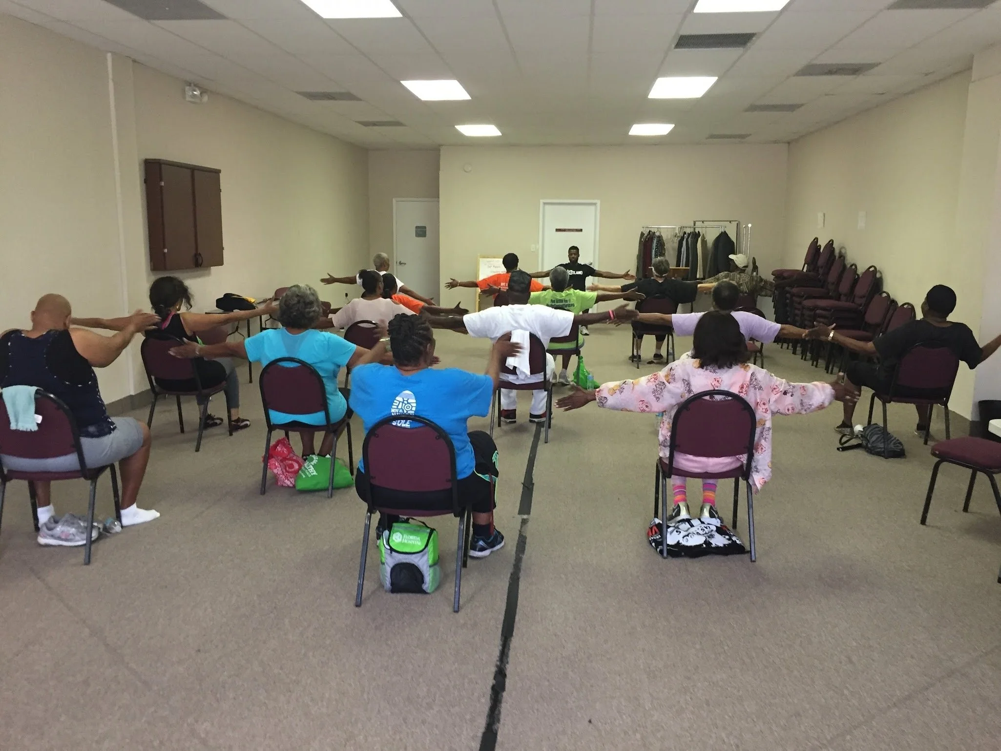 Group of diverse people participating in seated yoga or stretching exercise in a room with chairs and a coat rack.