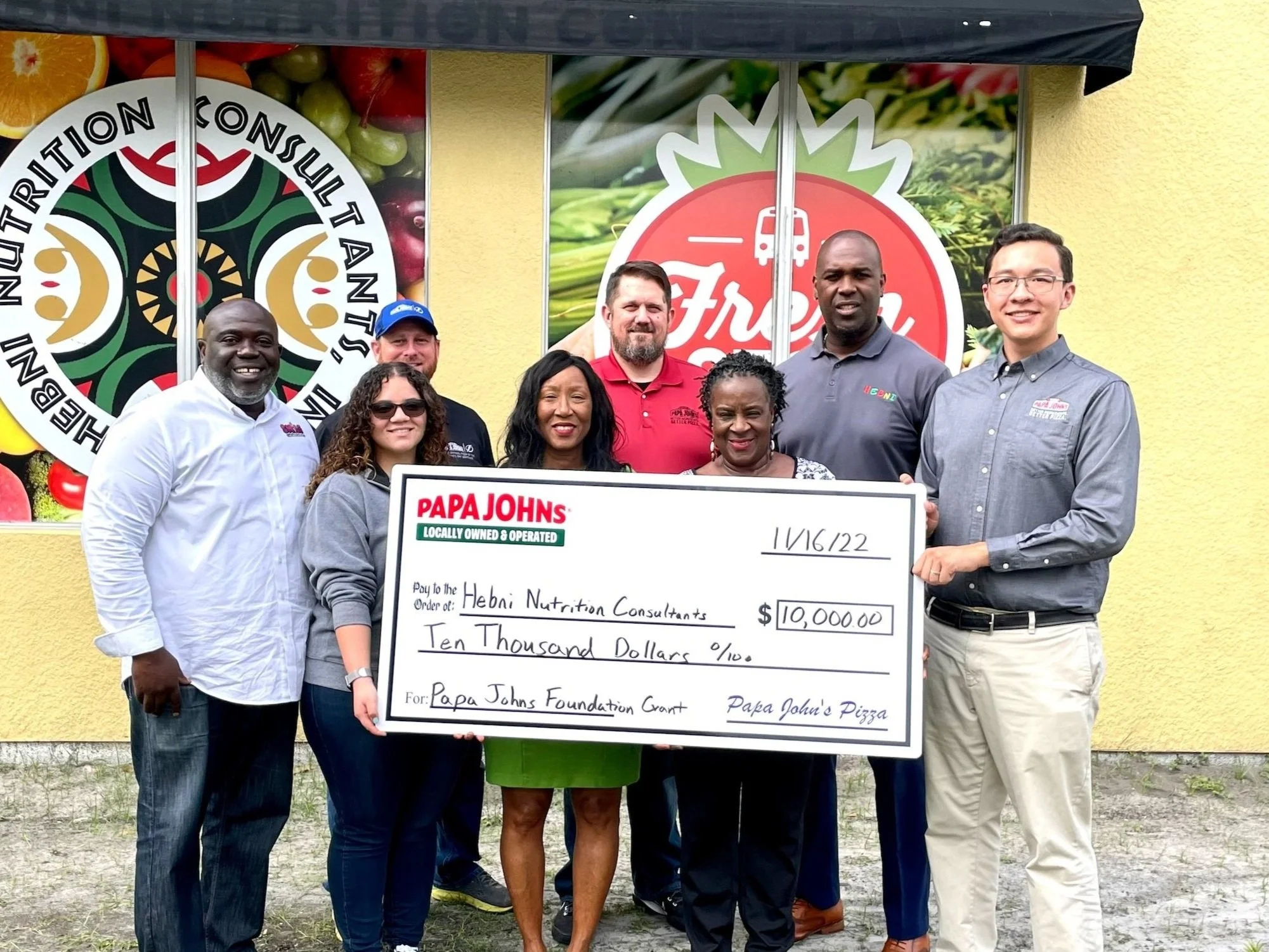 Group of eight people standing outside, holding a large check made out to Hebi Nutrition Consultants for ten thousand dollars, in front of a yellow building with large promotional signs for Fresh and a nutrition consultation brand.