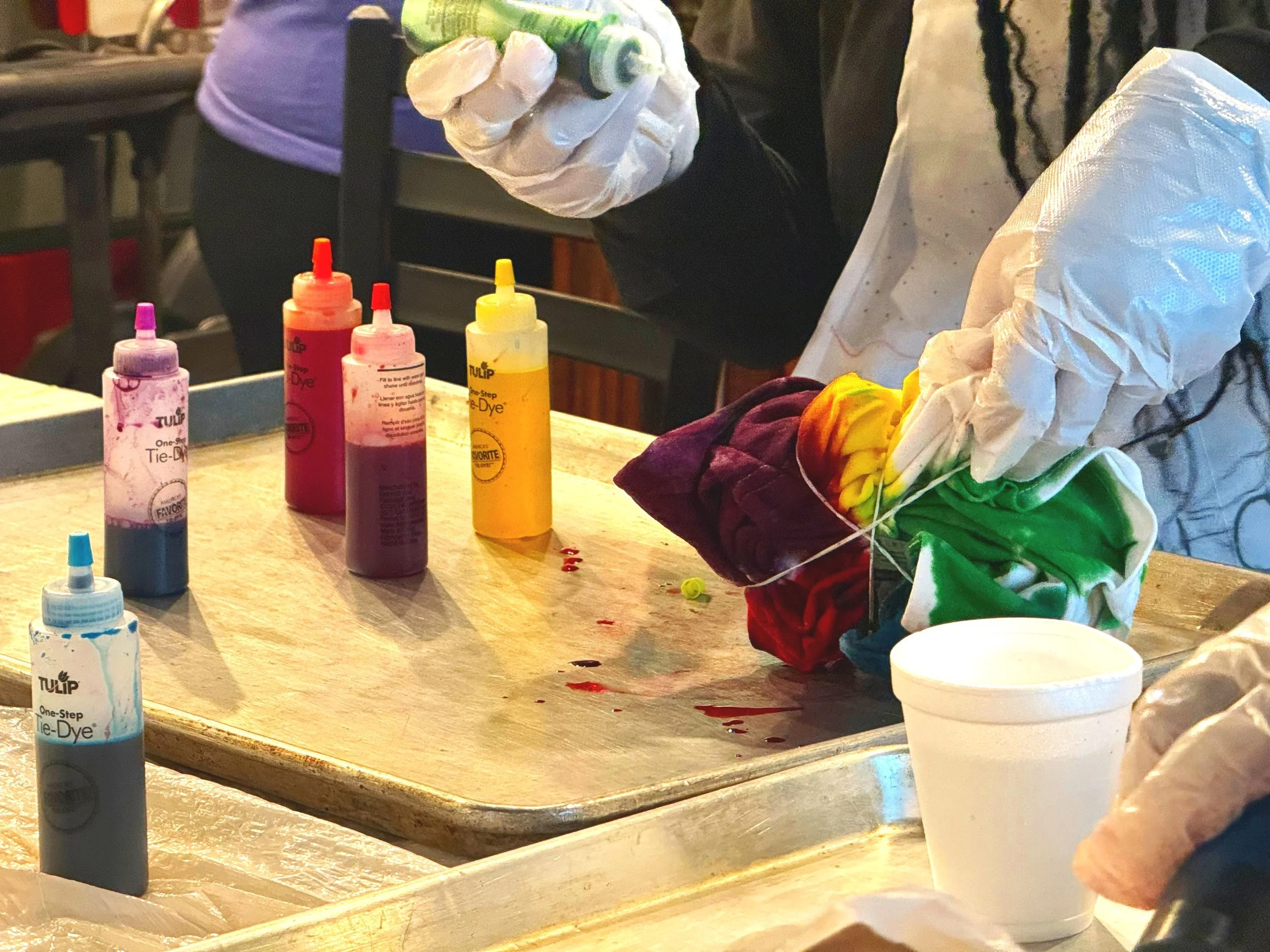 Person wearing gloves creating tie-dye fabric with bottles of colorful dye and a white container of water on a table.