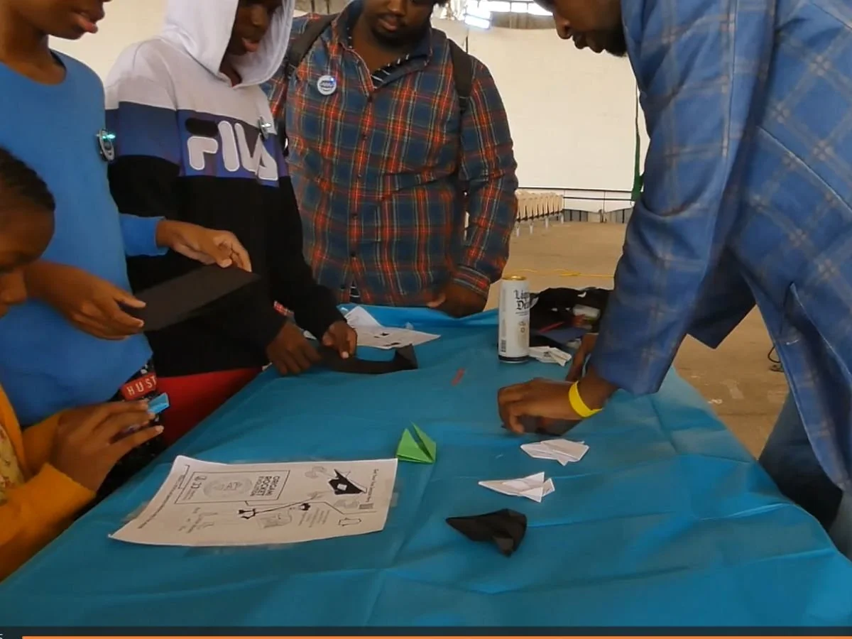 Group of people gathered around a blue table, folding paper and engaging in origami activity, with instructions and supplies on the table.