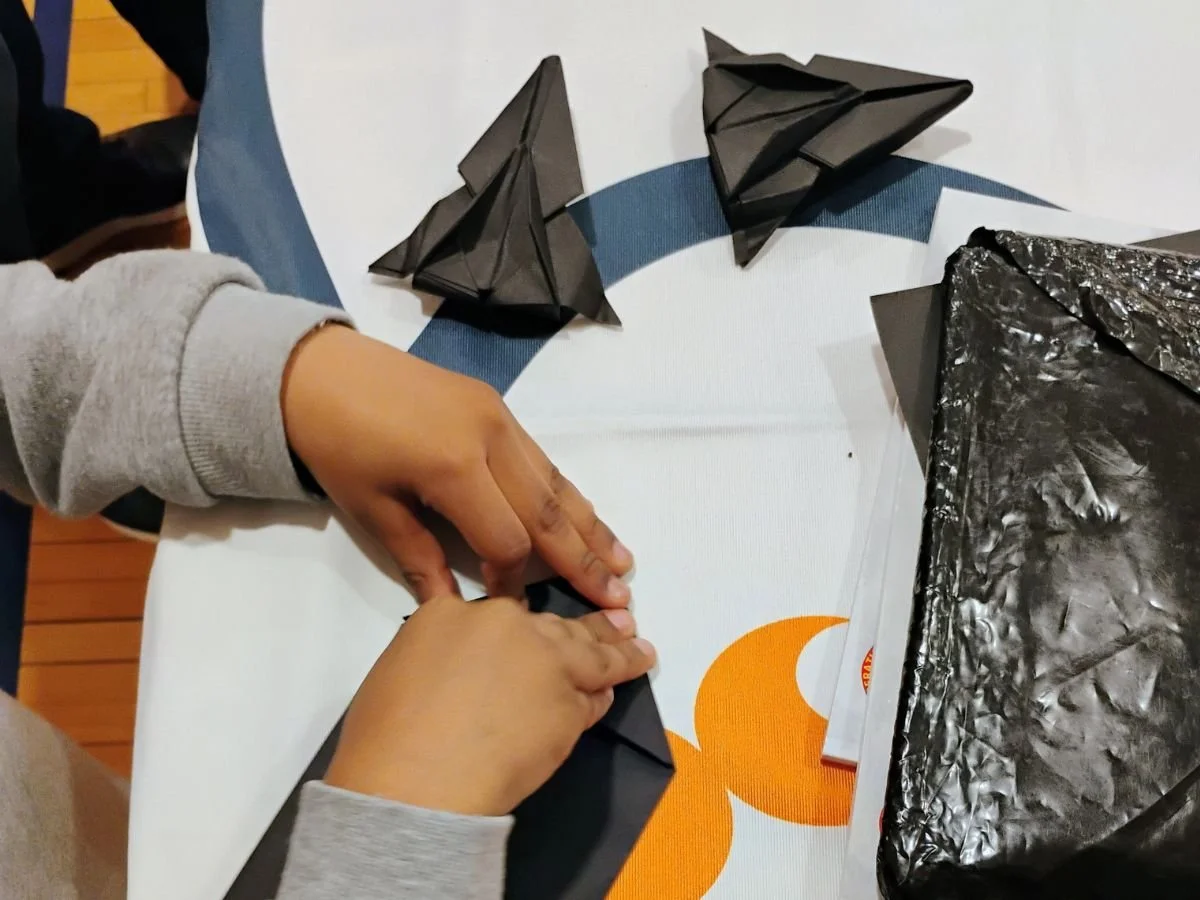 A person folding black origami paper models on a table that has a white tablecloth with an orange design, next to a black plastic bag.