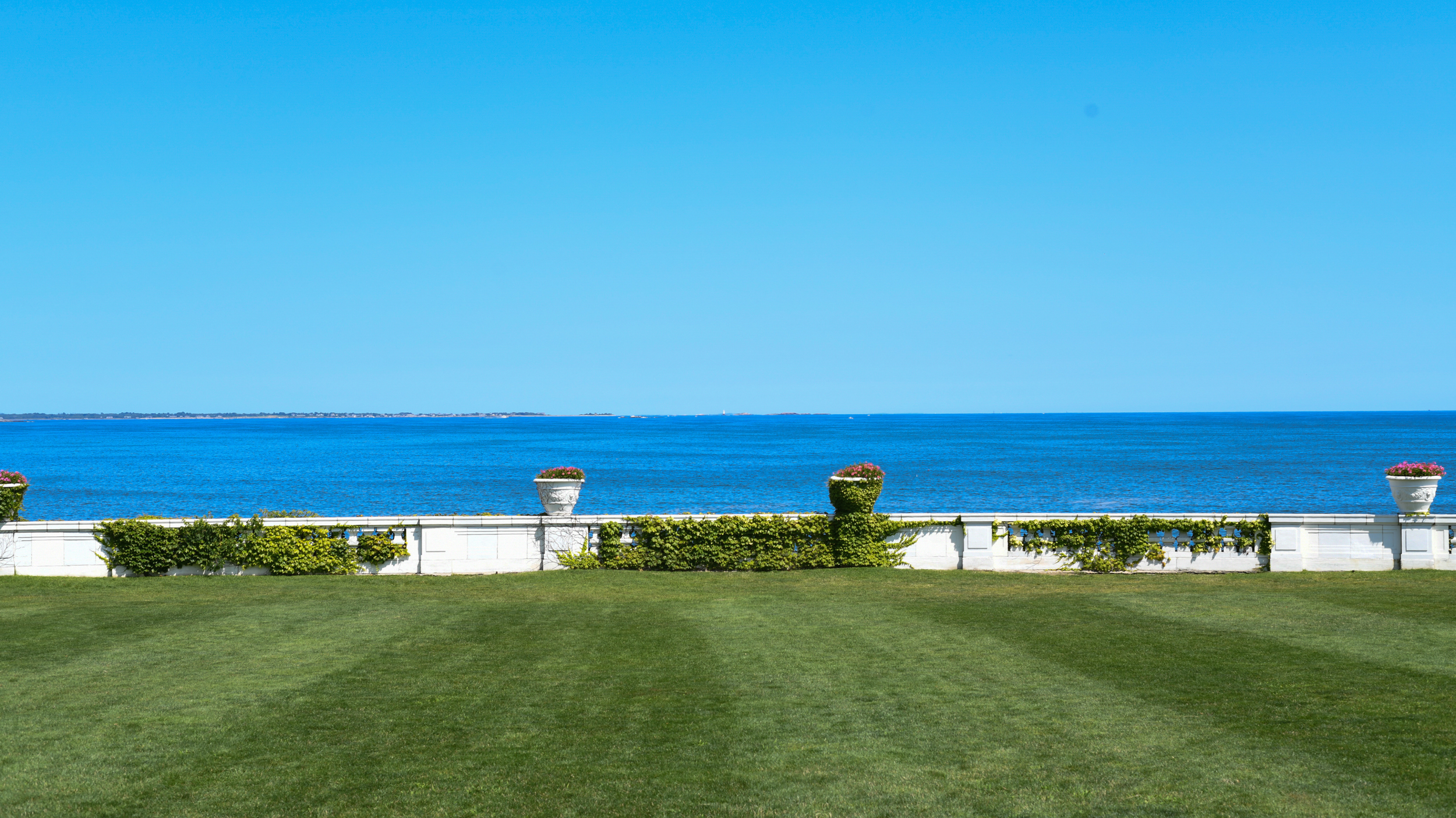 A view of the ocean with a bright blue sky, behind a white decorative wall with potted plants and green grass in the foreground.