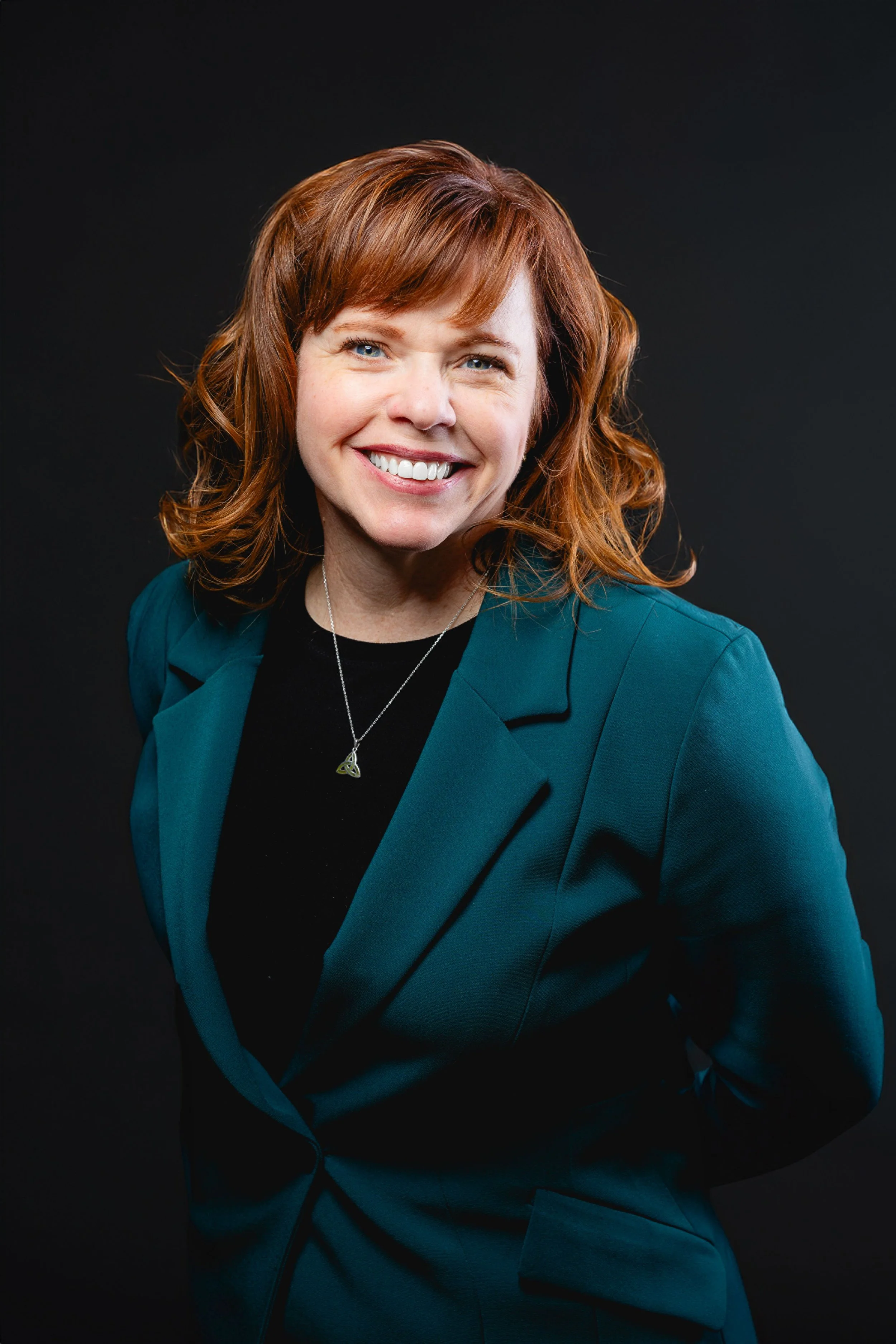 A woman with shoulder-length red hair, wearing a teal blazer over a black top, smiling at the camera against a dark background.