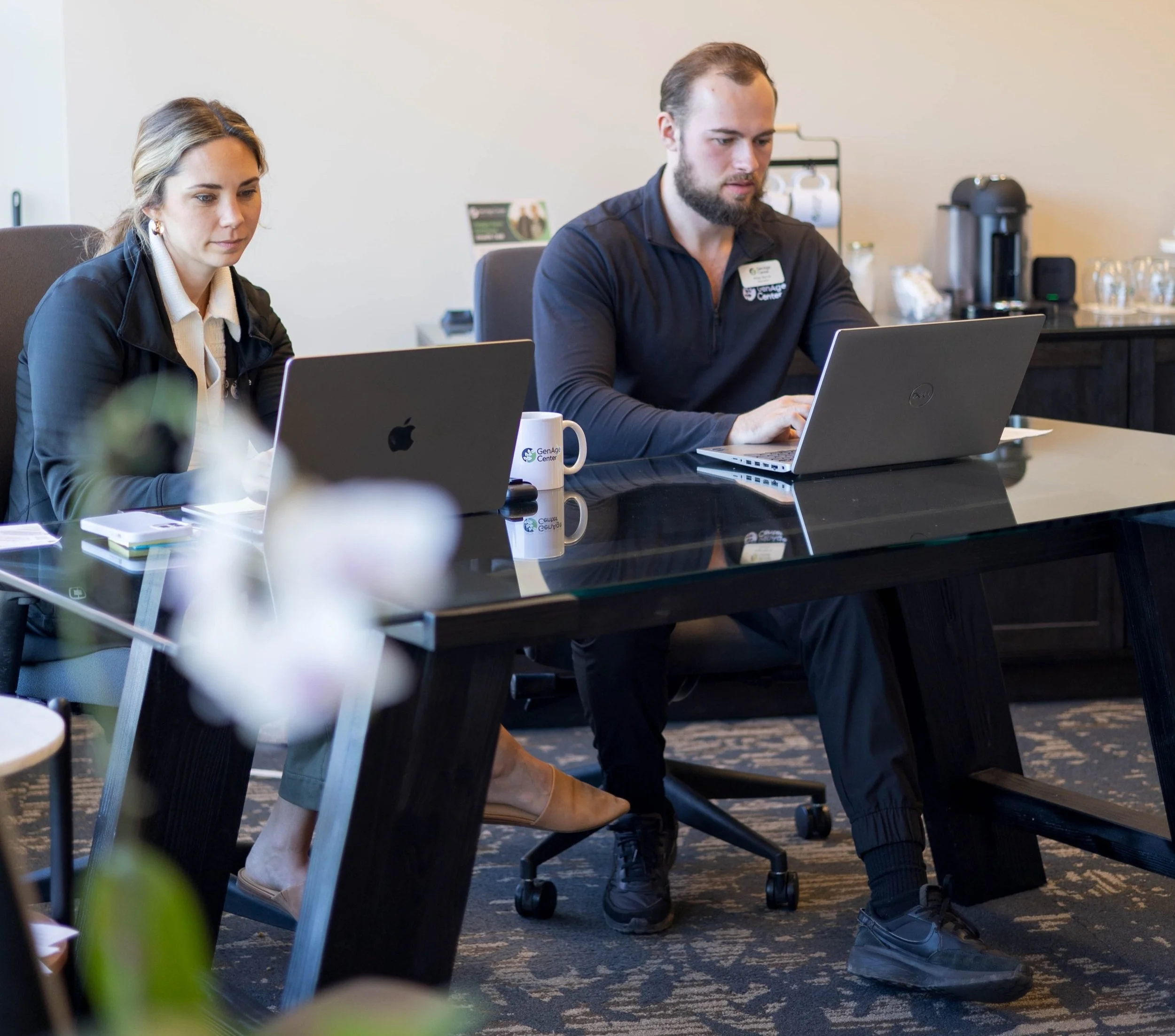 Two people working on laptops at a conference table, a woman on the left and a man on the right, in a professional office setting.