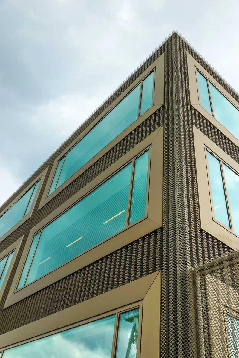 Modern kantoorgebouw met grote glazen ramen en metalen gevelbekleding, gezien vanaf een hoek onder een bewolkte hemel.