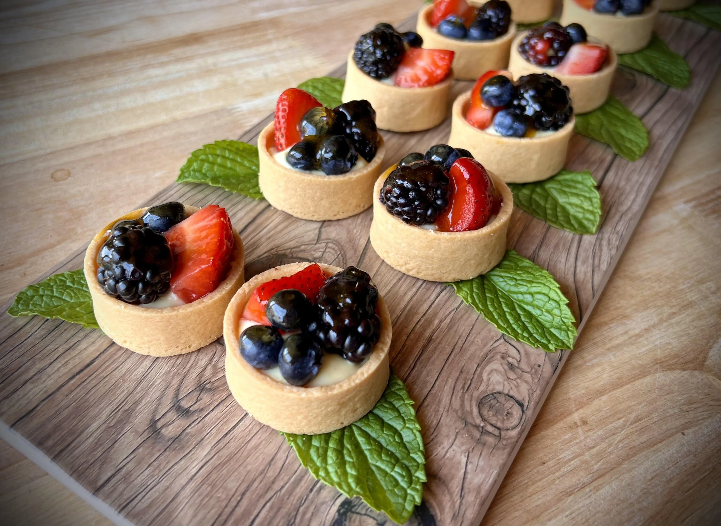 Mini fruit tartlets on a wooden board, topped with strawberries, blackberries, blueberries, and mint leaves.