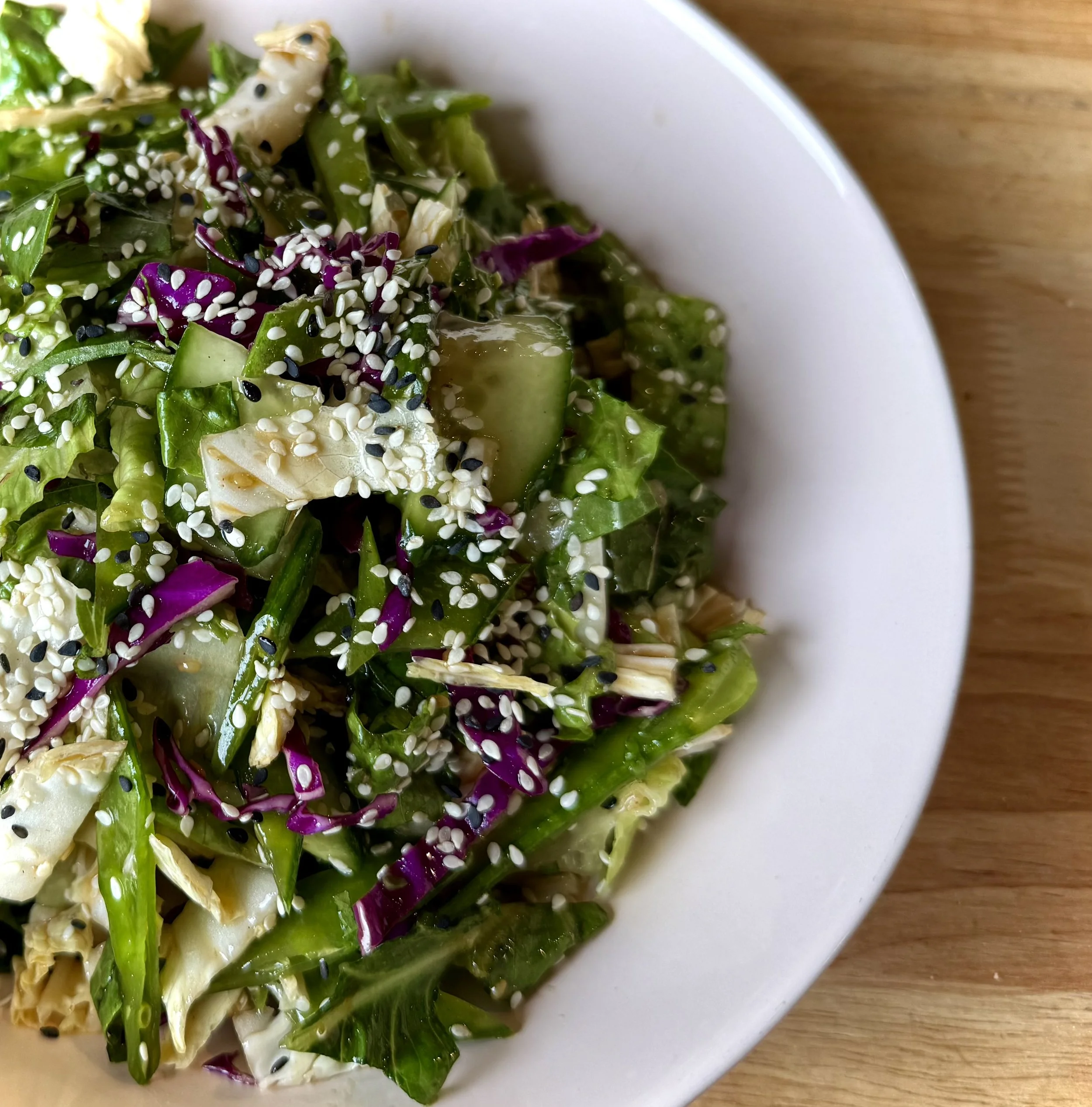 Close-up of a vegetable salad with cucumbers, purple cabbage, lettuce, and shredded chicken, topped with white and black sesame seeds in a white bowl on a wooden surface.